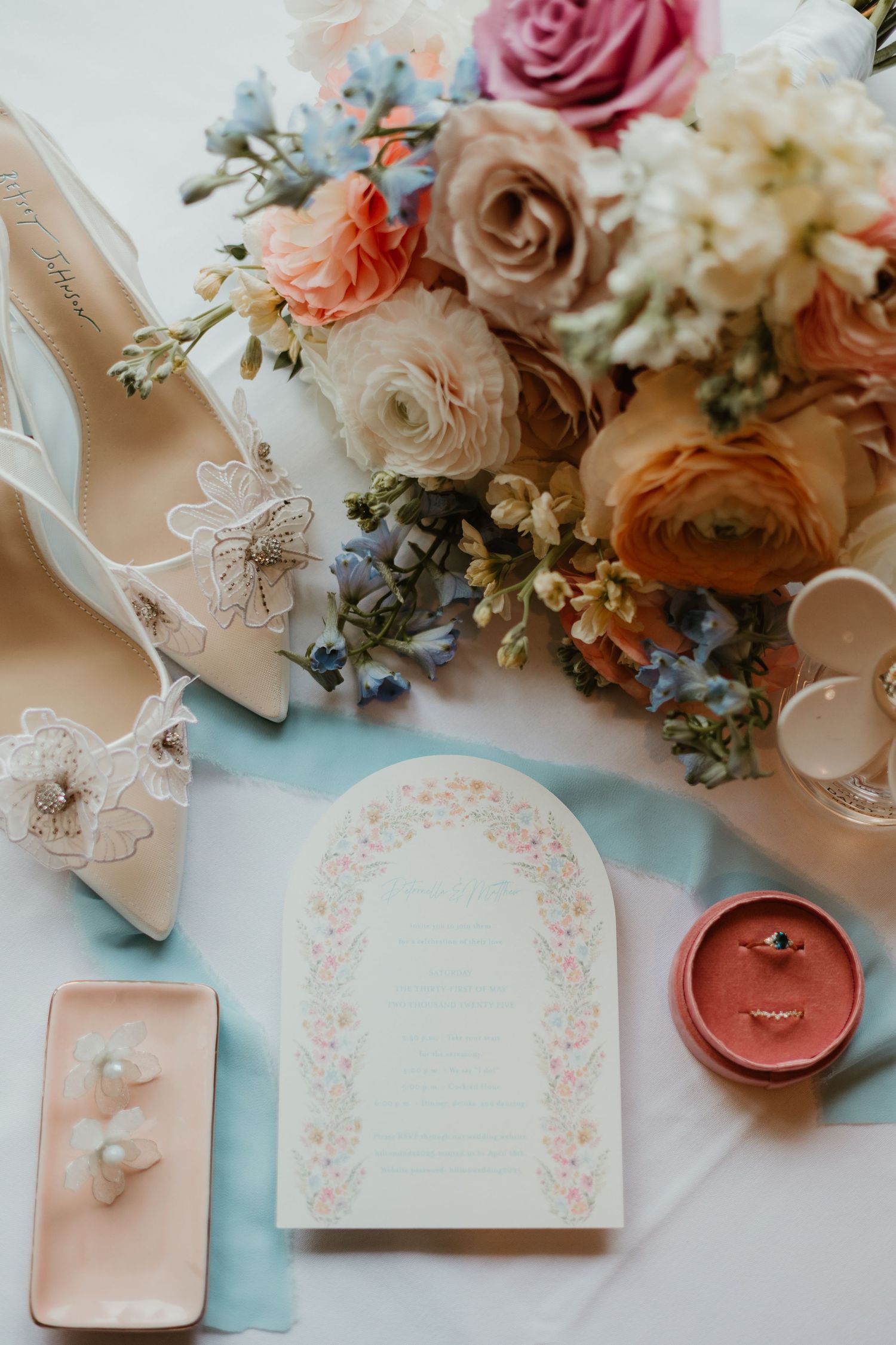 Wedding flat lay featuring bridal heels, floral bouquet, invitation card, pearl earrings, and diamond ring on blue ribbon.