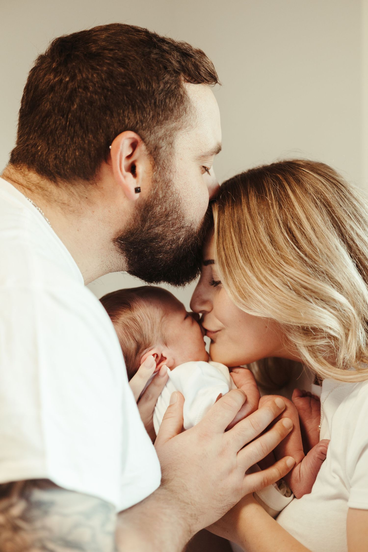 New parents share a kiss while holding their sleeping newborn baby together.