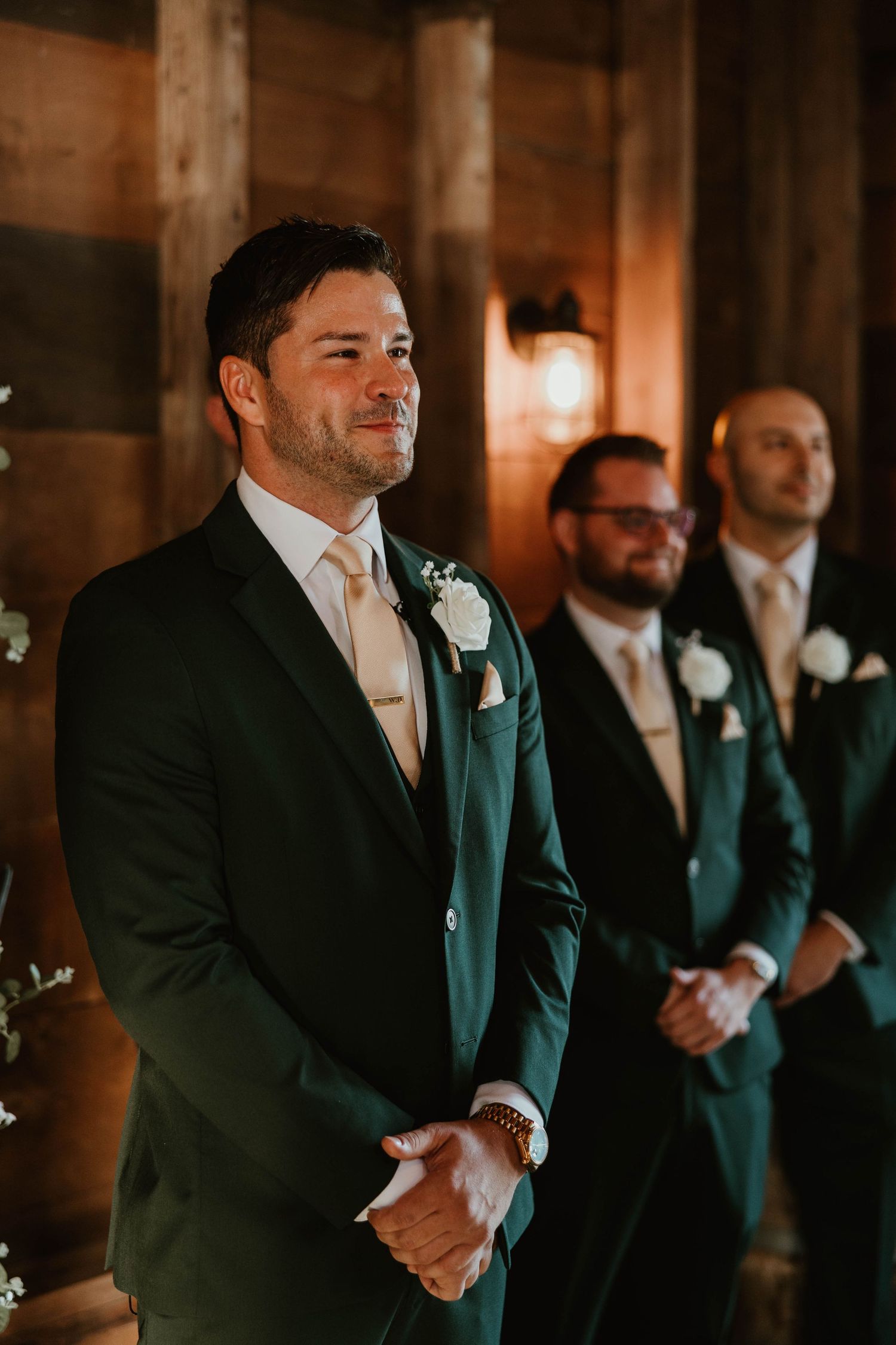 Emotional groom in green suit watches bride walk down aisle flanked by groomsmen in barn wedding.