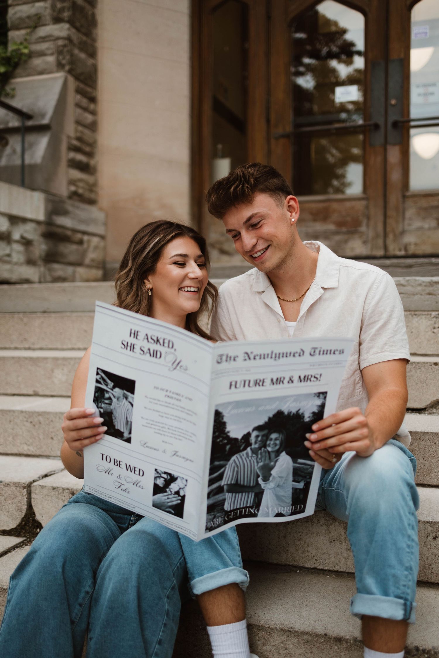 Smiling engaged couple sitting on stone steps reading The Newlywed Times engagement announcement newspaper.