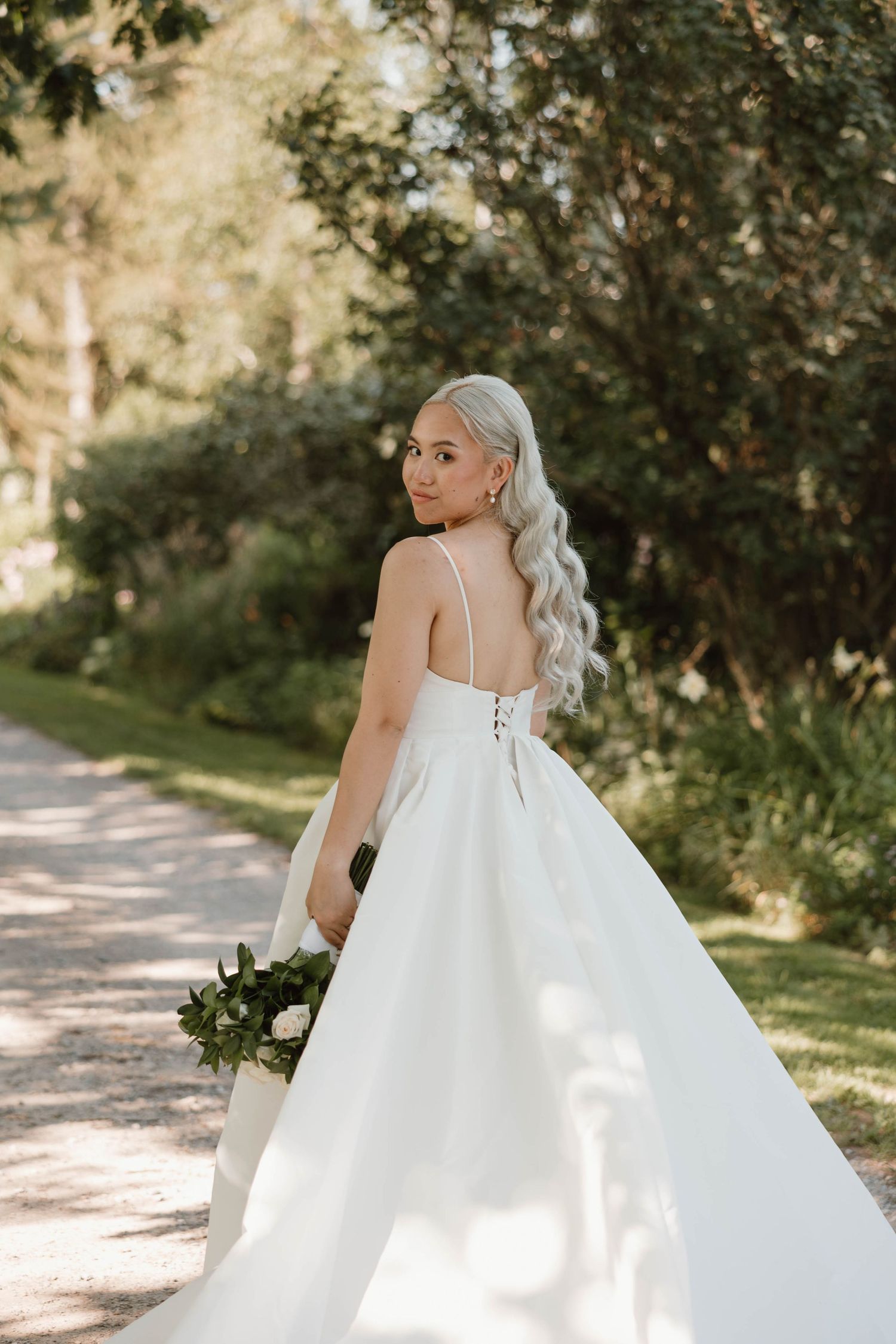 Bride in white ball gown with lace-up back holding bouquet on garden path.