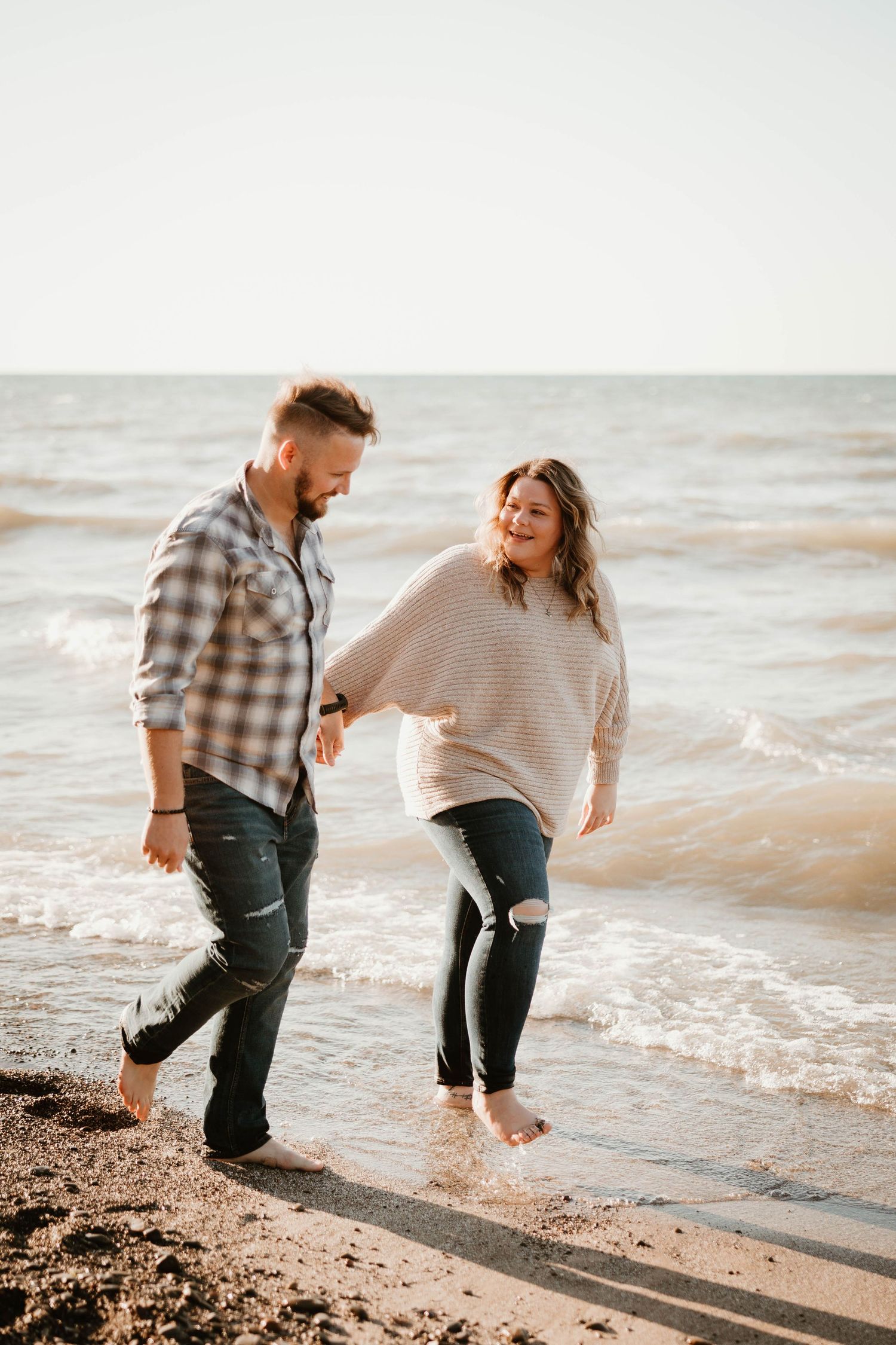 Couple laughing and holding hands while walking barefoot along a sandy beach shoreline.