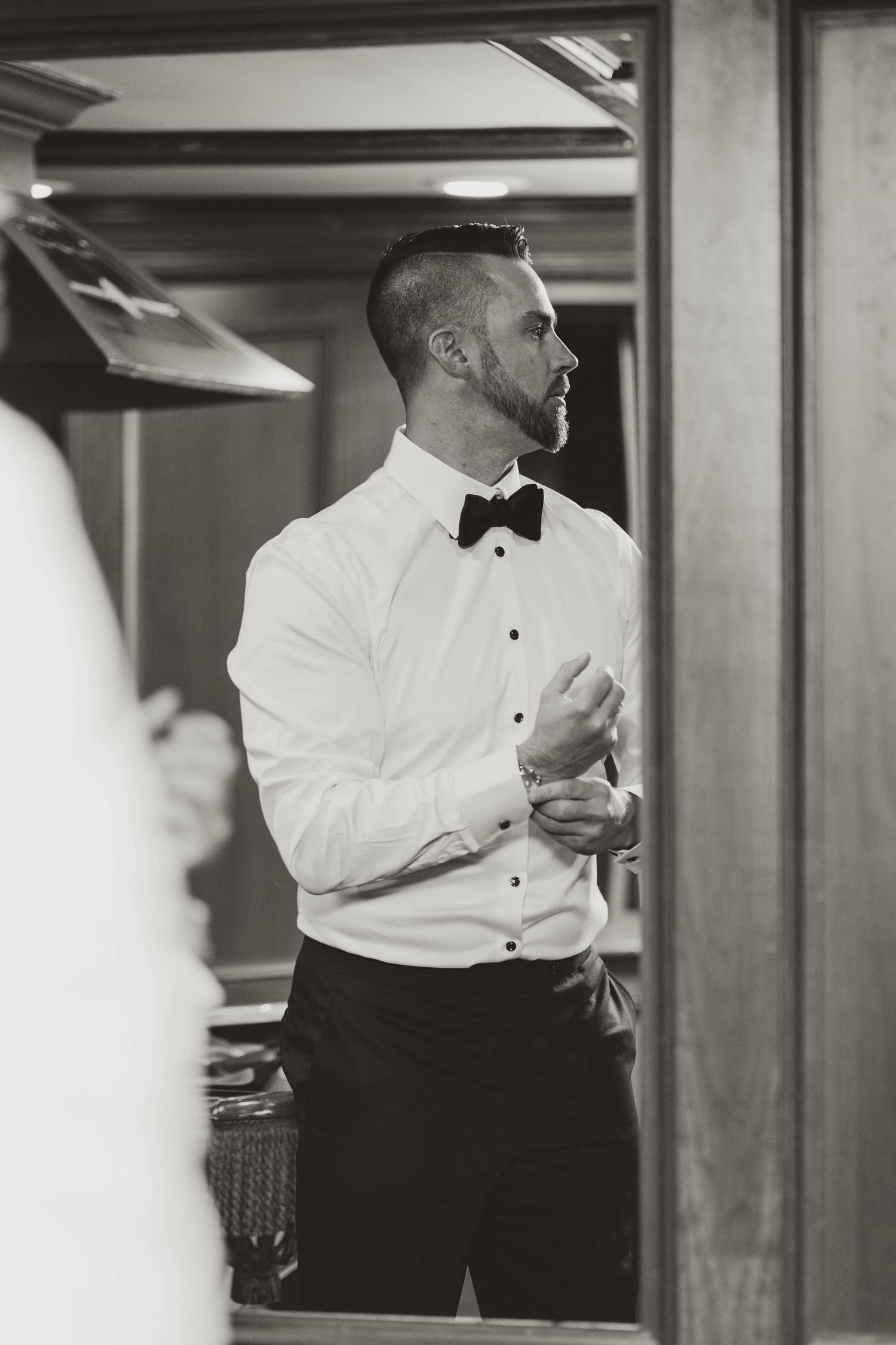 Black and white portrait of groom in tuxedo adjusting cufflinks before wedding.