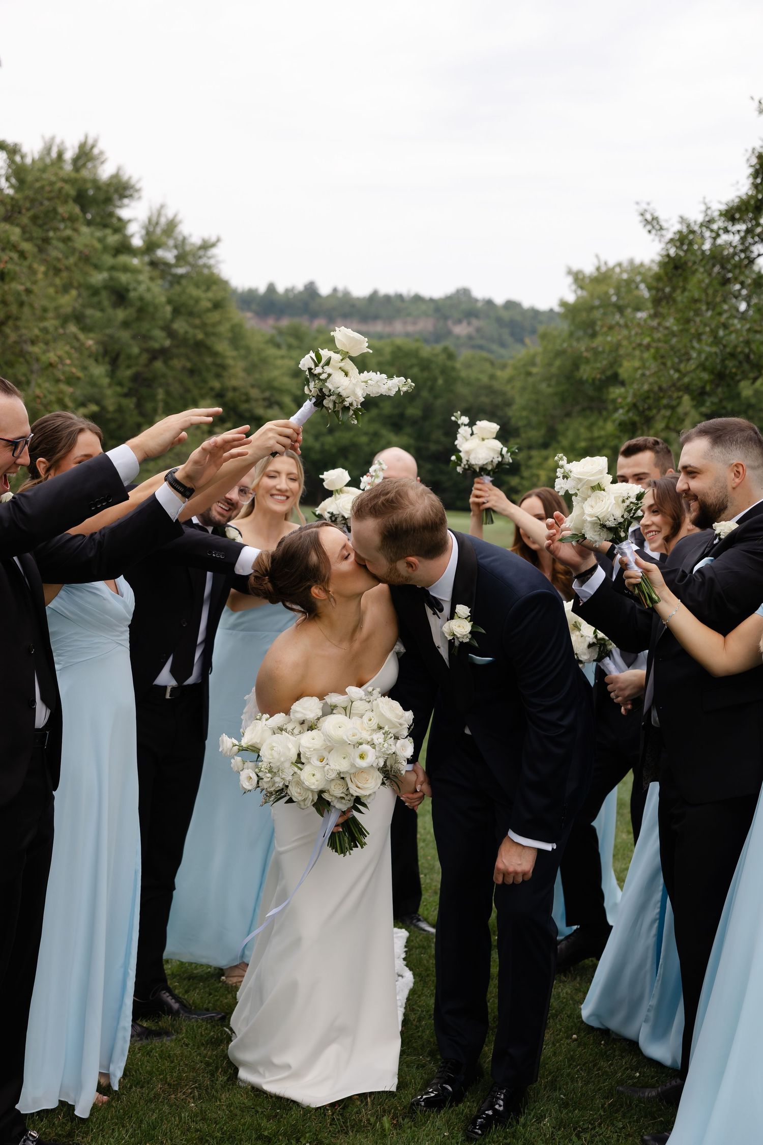 Bride and groom share a kiss surrounded by wedding party in light blue dresses and dark suits holding white bouquets outdoors.