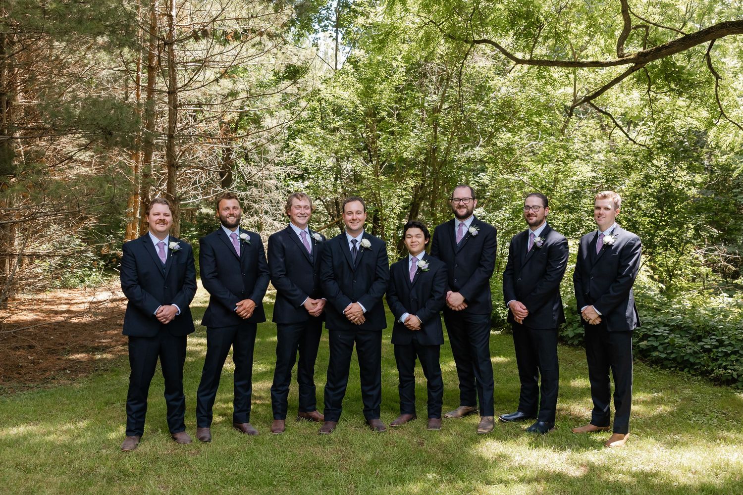 Groom poses with seven groomsmen in navy suits among tall trees on wedding day.