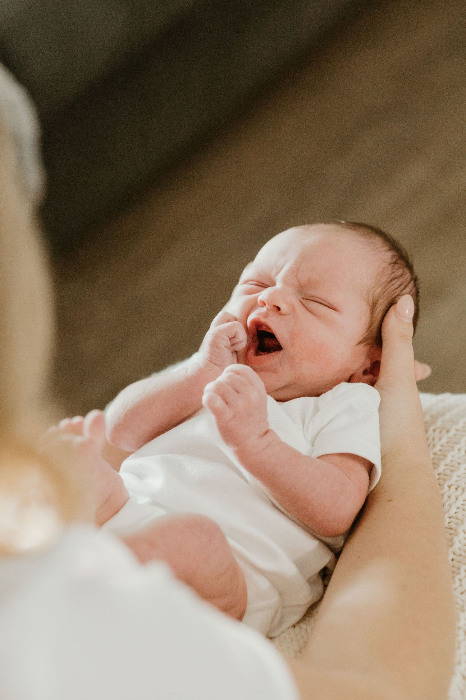 Newborn baby yawning while being cradled in mothers arms in white onesie.