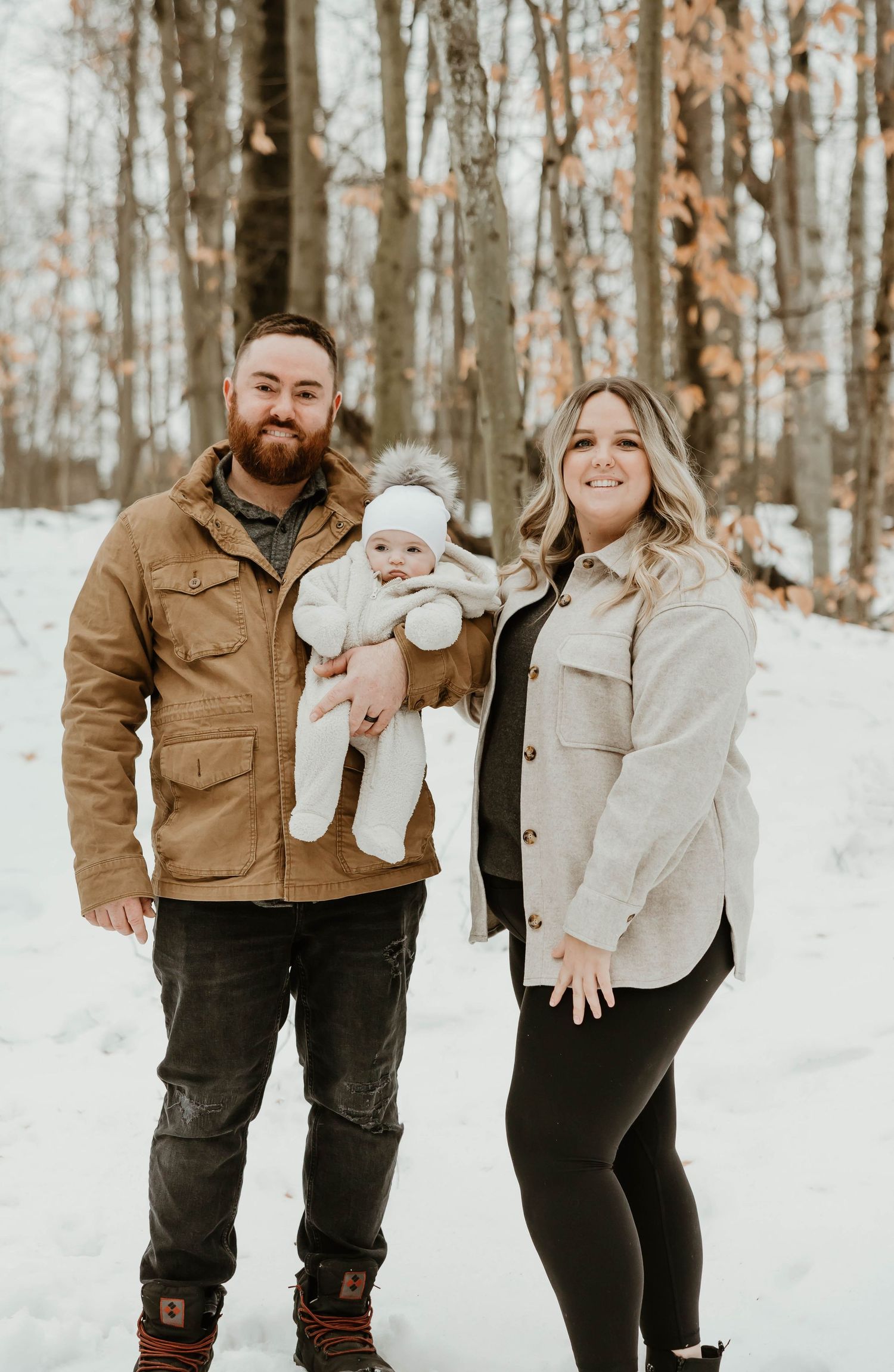 Couple standing in snowy forest holding baby dressed in white winter outfit.