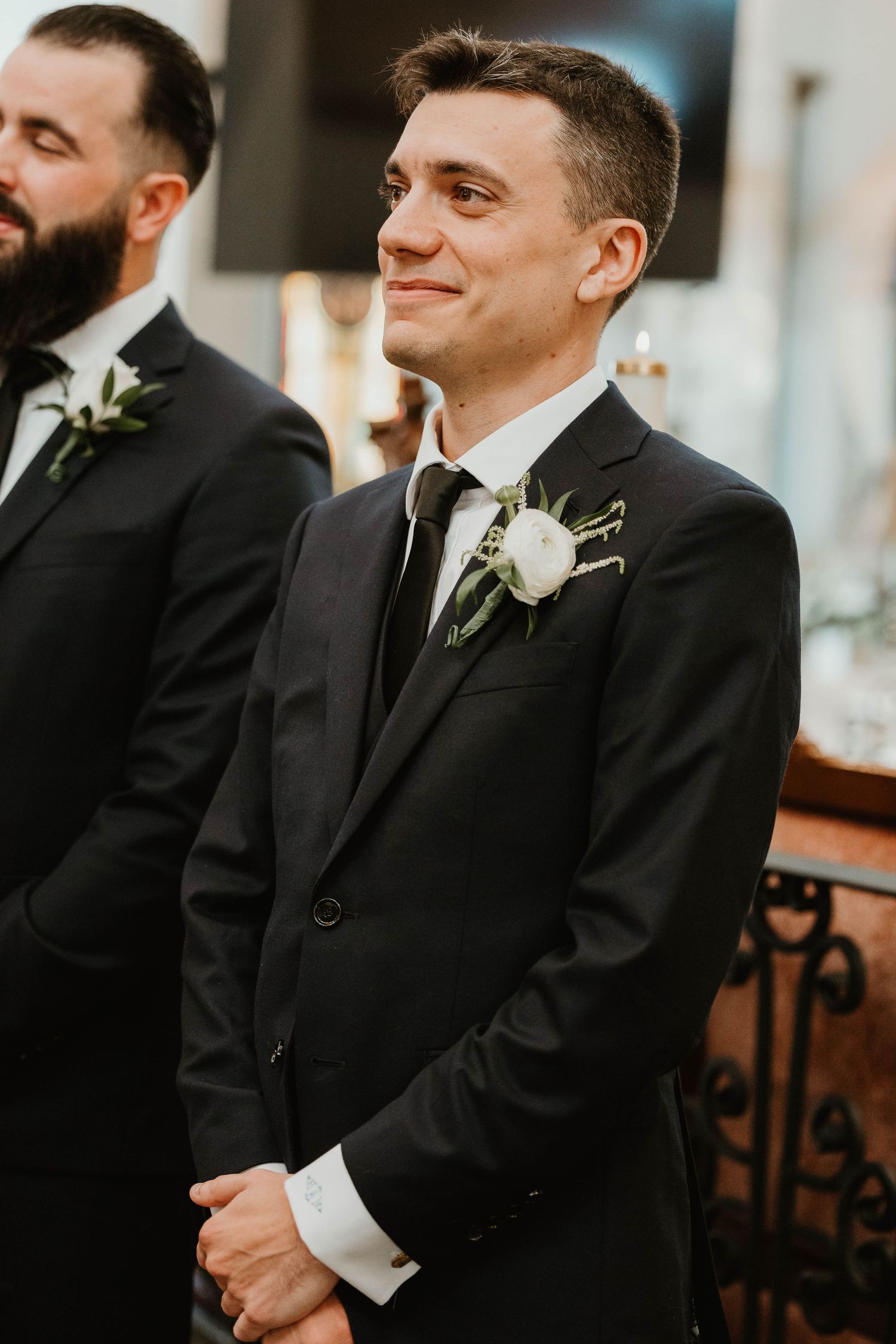 Smiling groom in black suit with white rose boutonniere awaits bride at church wedding ceremony altar.
