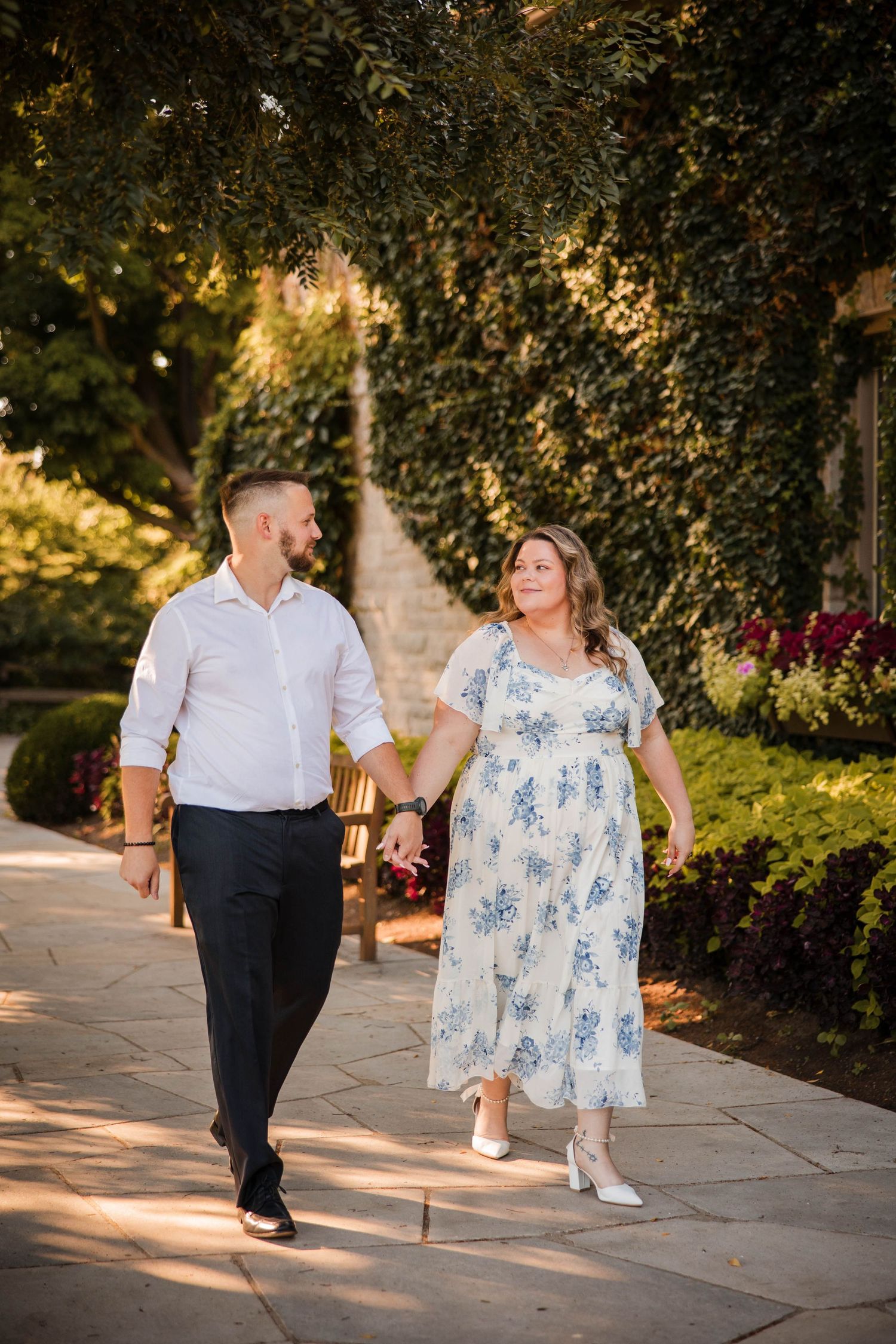 Couple holding hands and walking along a sunlit garden path surrounded by greenery.