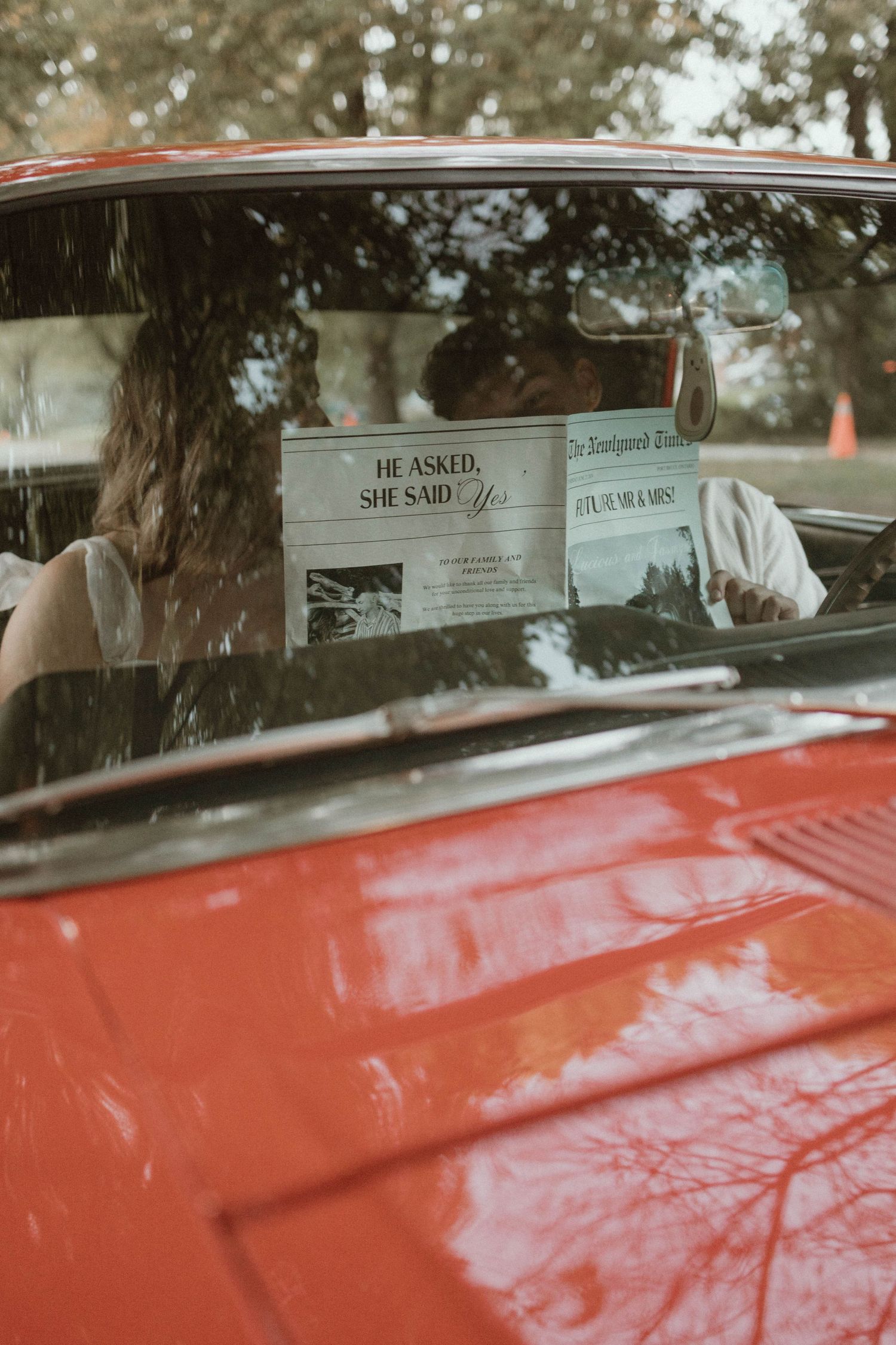 Couple kissing in a red vintage car holding a newspaper headline reading He Asked She Said Yes.