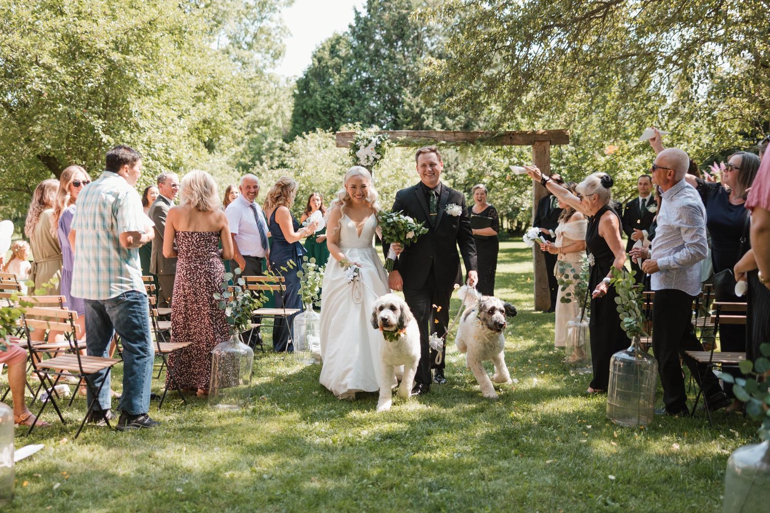 Newlyweds walking down outdoor aisle with two large white dogs after ceremony.