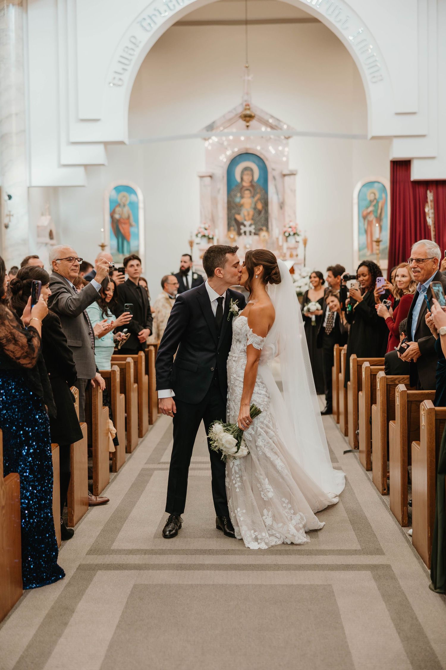 Newlyweds share first kiss walking down church aisle as guests celebrate with applause.