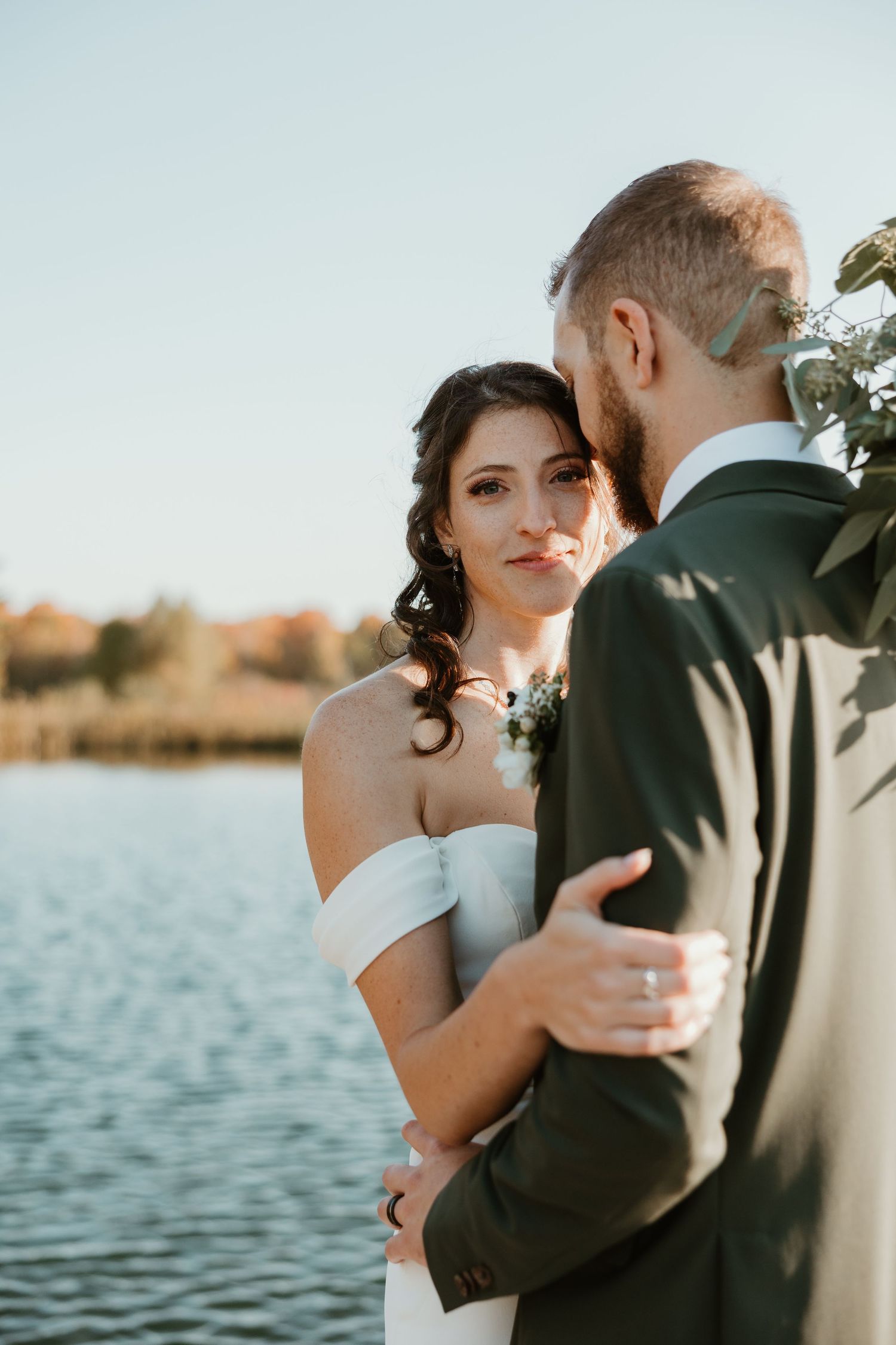 Bride in off-shoulder gown rests head on groom's shoulder near a sunlit lake outdoors.