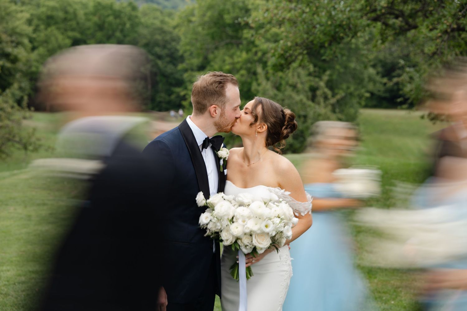 A bride and groom share a romantic kiss at their outdoor wedding, surrounded by blurred guests in motion.