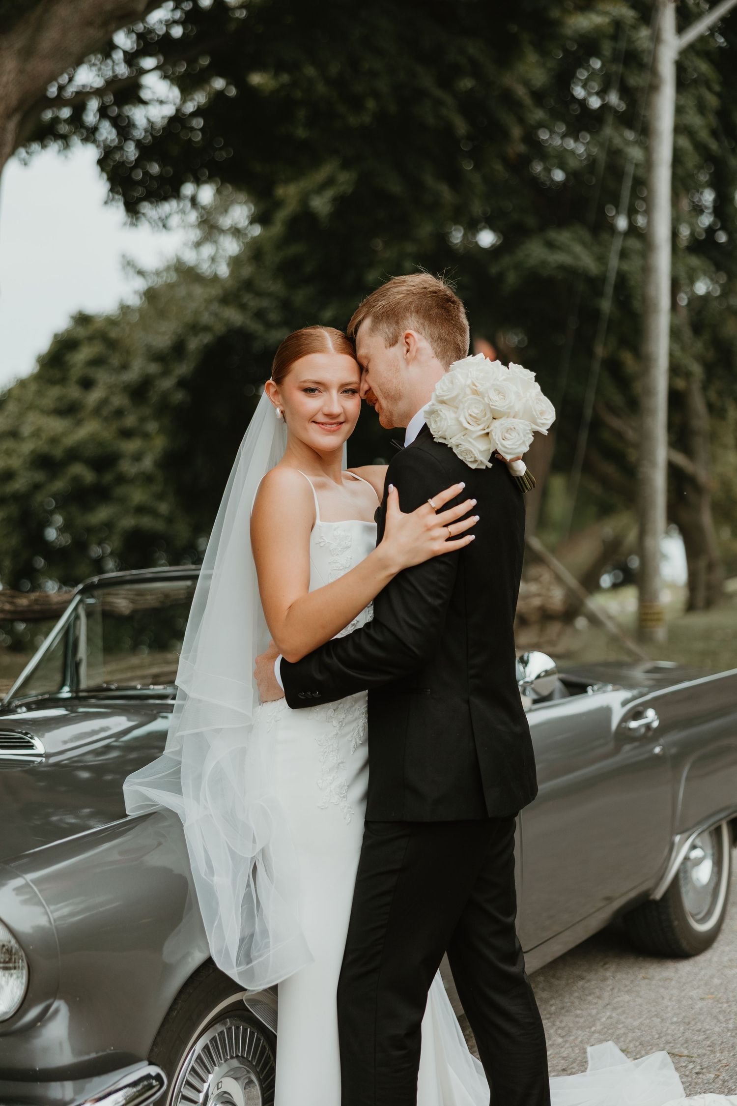 Bride and groom embrace romantically in front of a classic gray vintage convertible car outdoors.