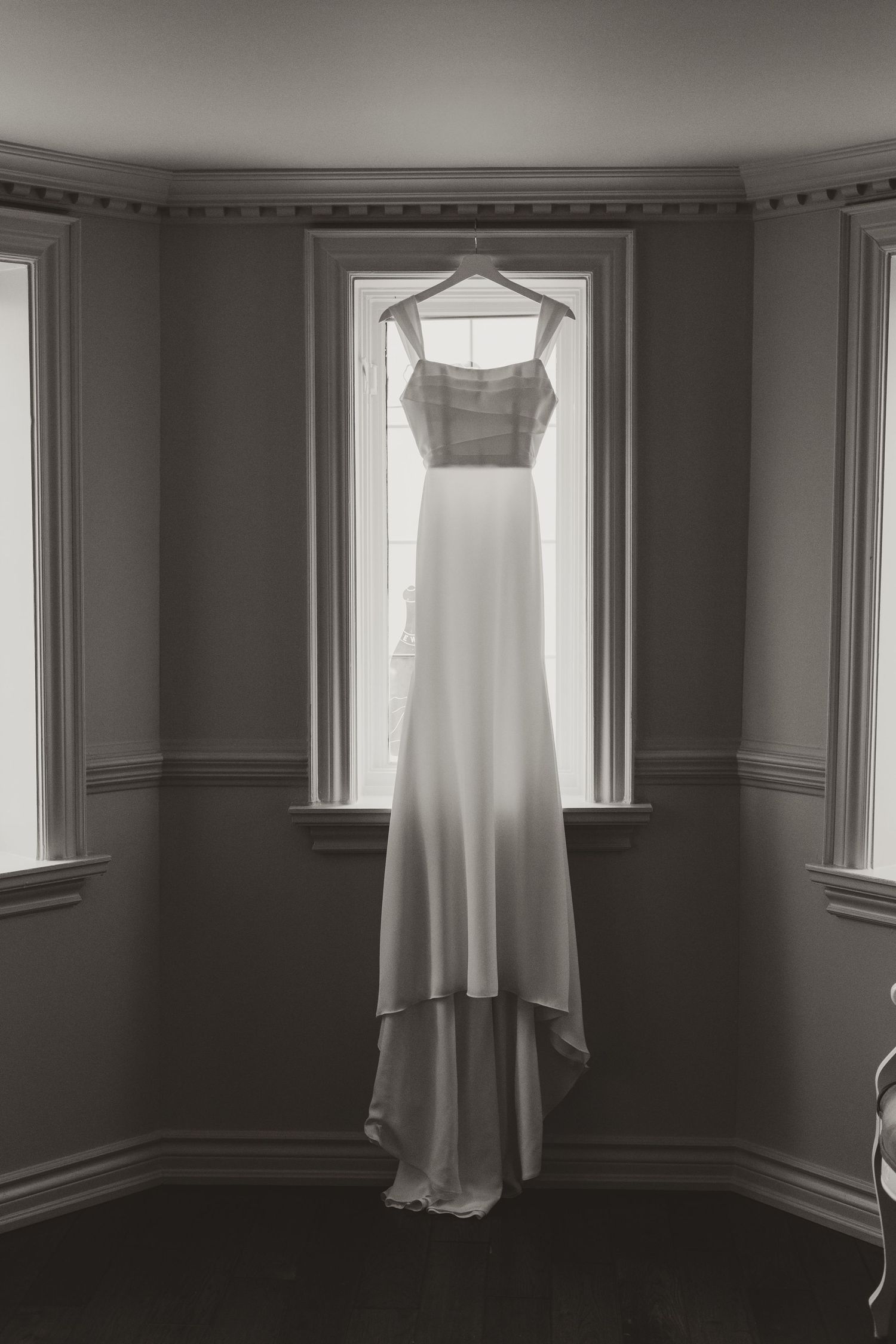 Elegant white bridal gown hanging on a hanger in a bay window in a black and white photo.