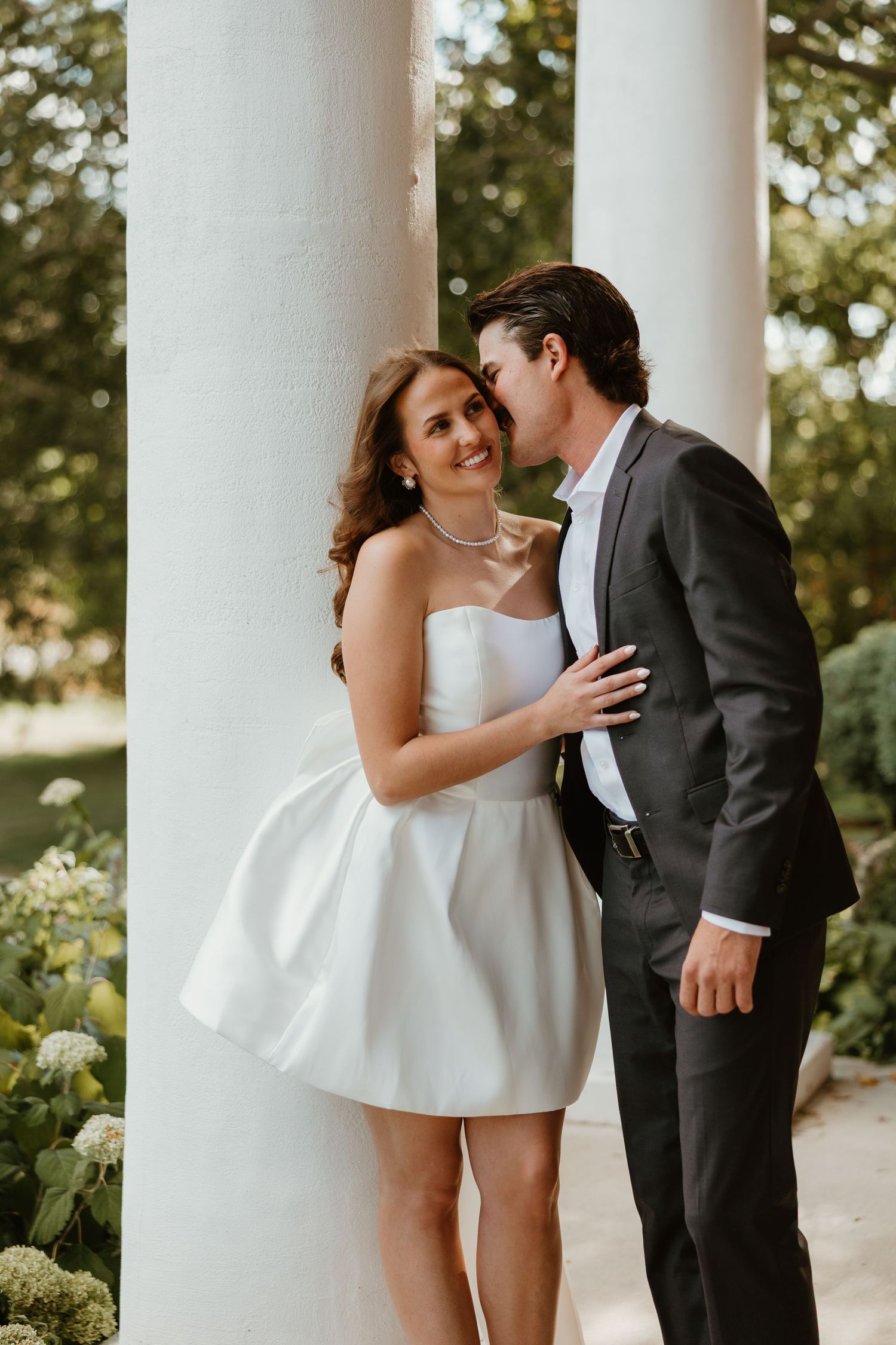 Couple sharing a romantic moment by white columns, bride in short white strapless dress, groom in dark suit.