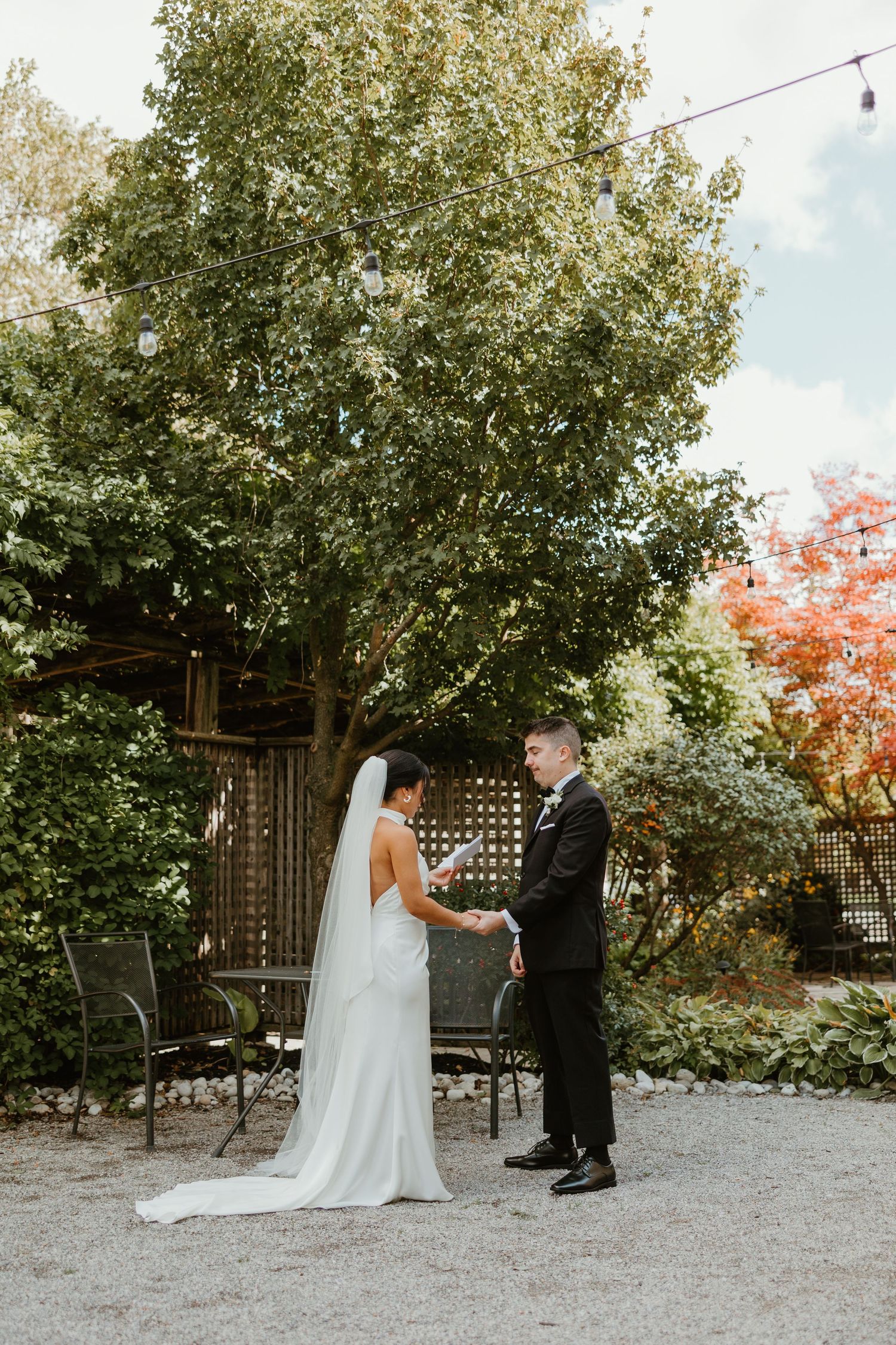 Bride in white gown with veil and groom in black suit hold hands in lush garden during wedding portraits.