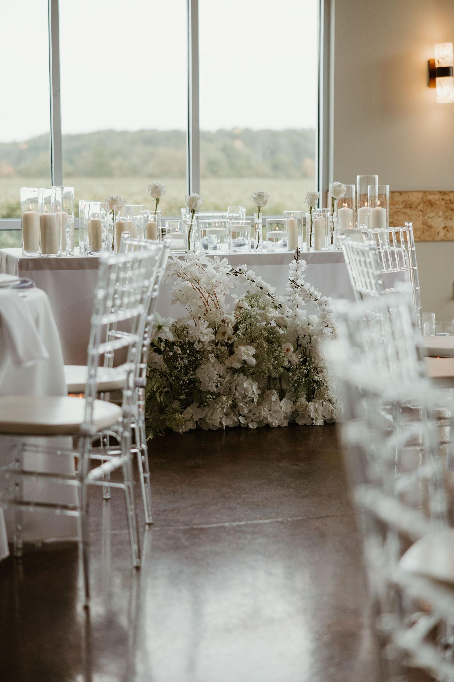 Elegant wedding reception hall with white floral centerpieces and ghost chairs by windows.
