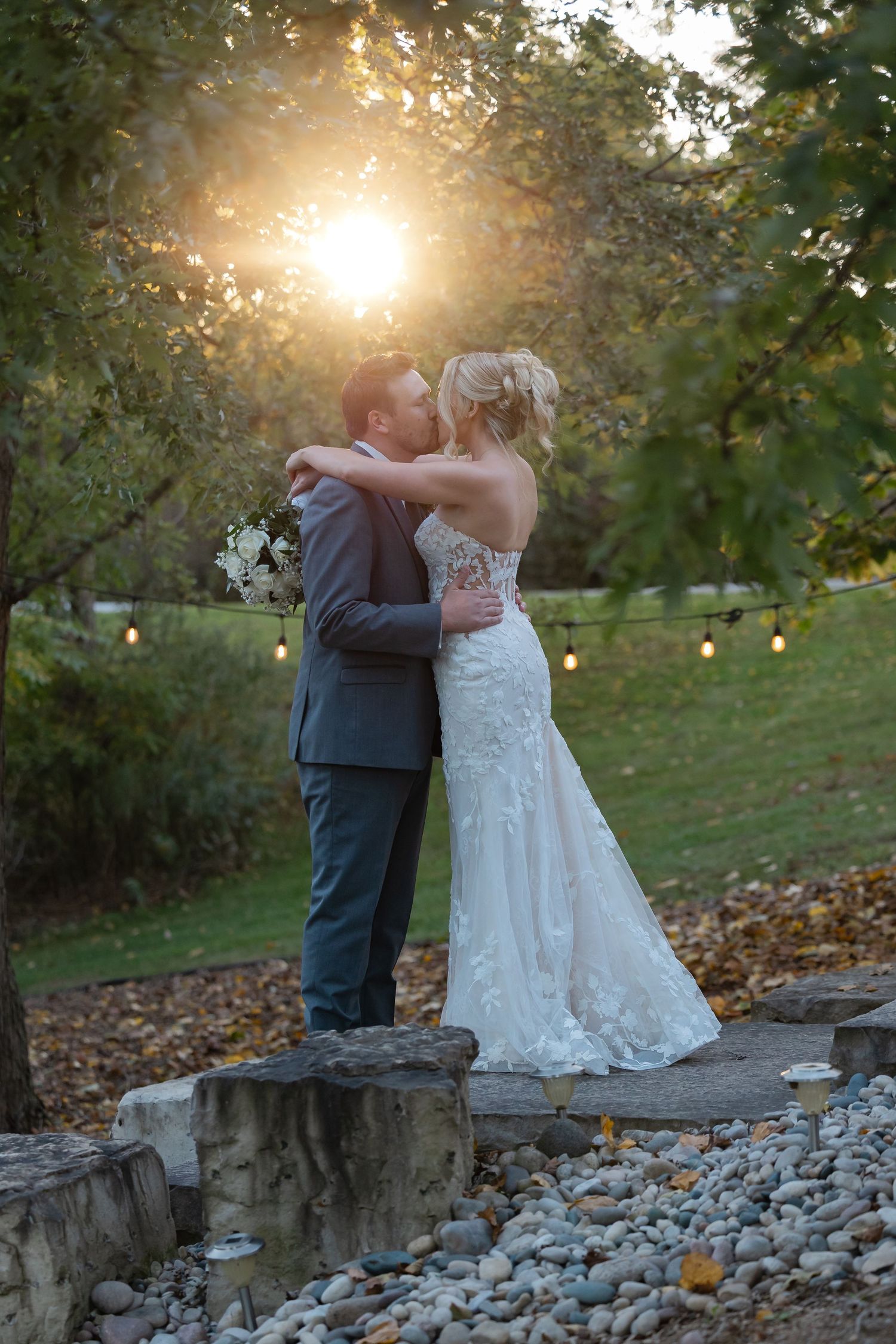 Bride in a lace wedding gown and groom in a gray suit embrace at sunset beside a stone ledge, with string lights and autumn leaves behind.