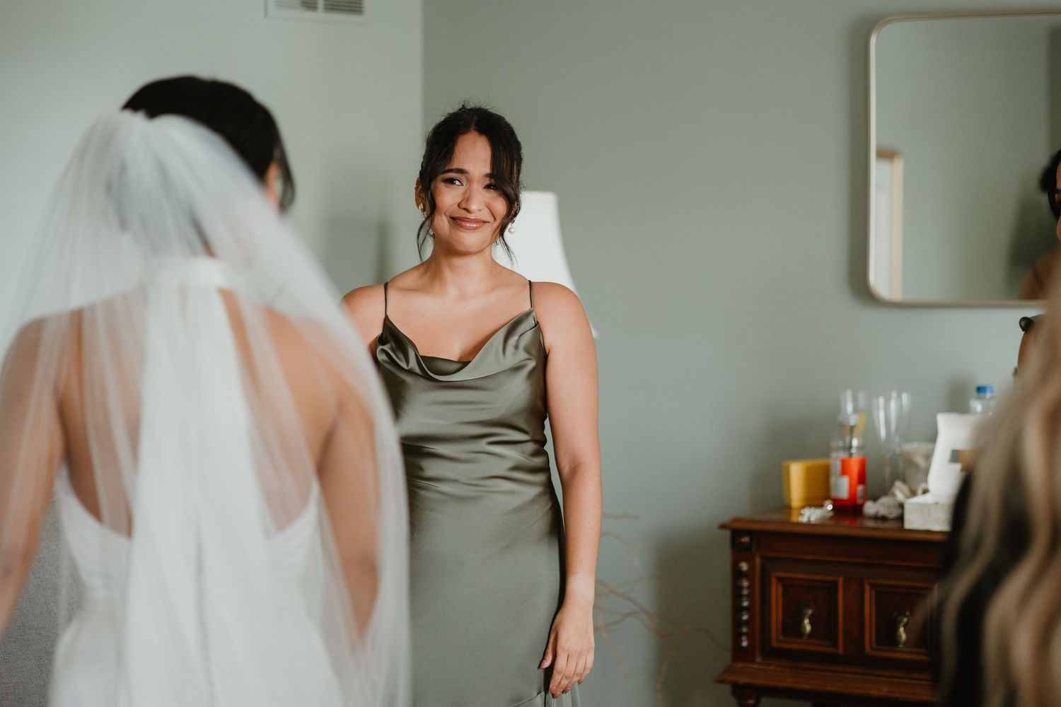 Bridesmaid in sage green satin dress smiles warmly at bride in white gown and veil during wedding preparations.