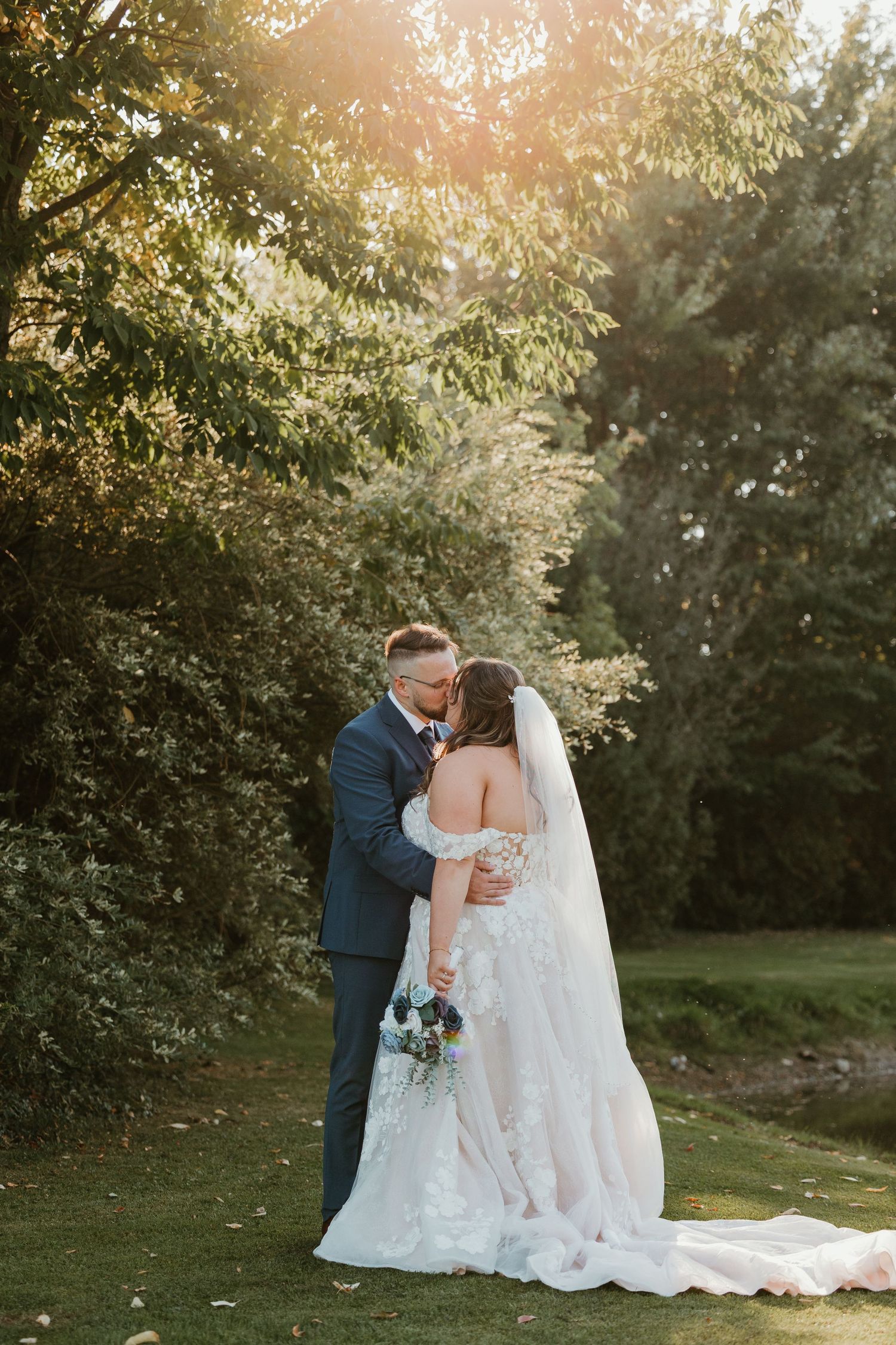 Bride and groom sharing a kiss under sunlit trees in a garden during golden hour wedding photos.