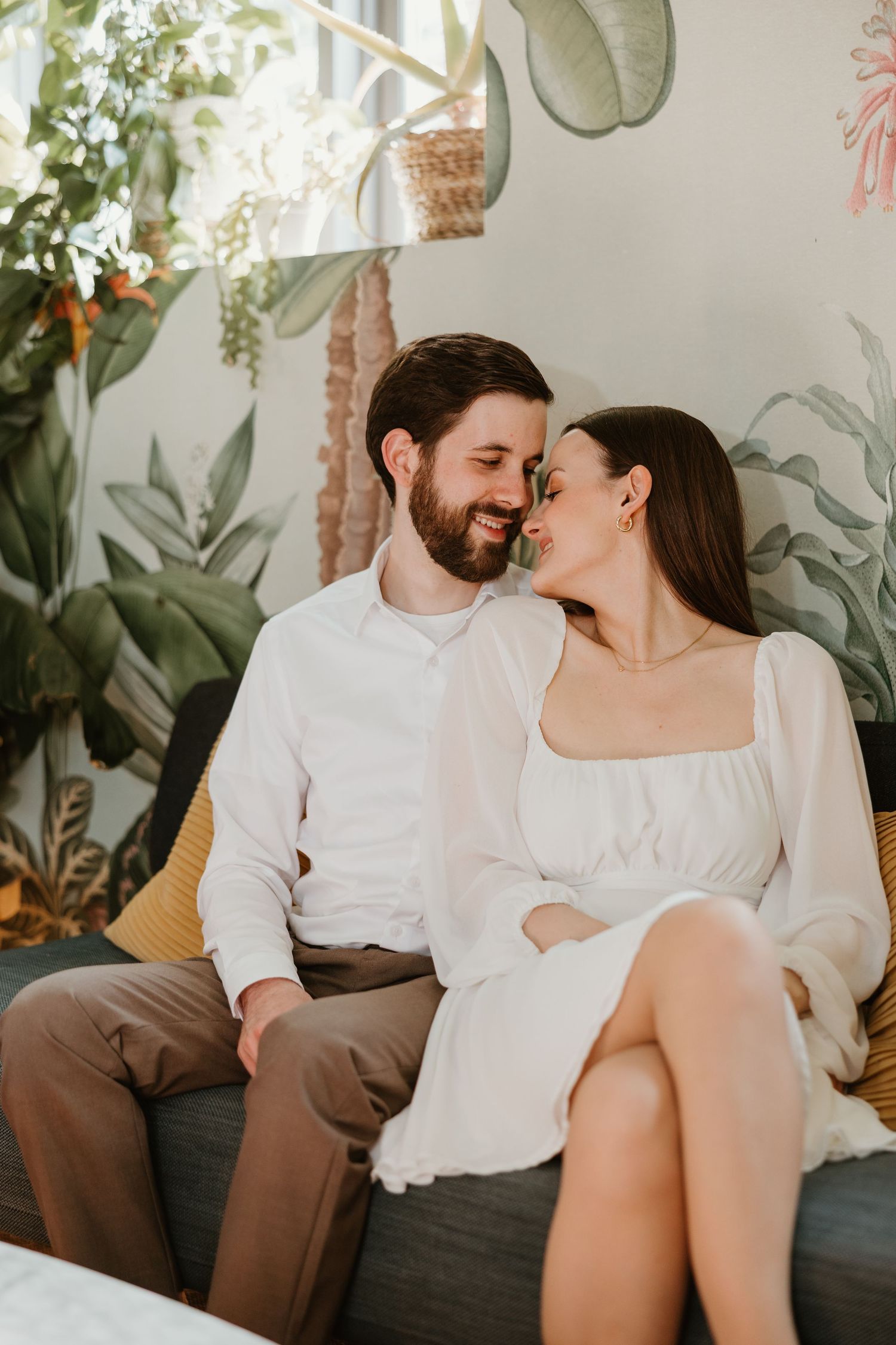 Couple in white outfits sitting close together sharing a romantic moment on a couch with tropical botanical wallpaper.