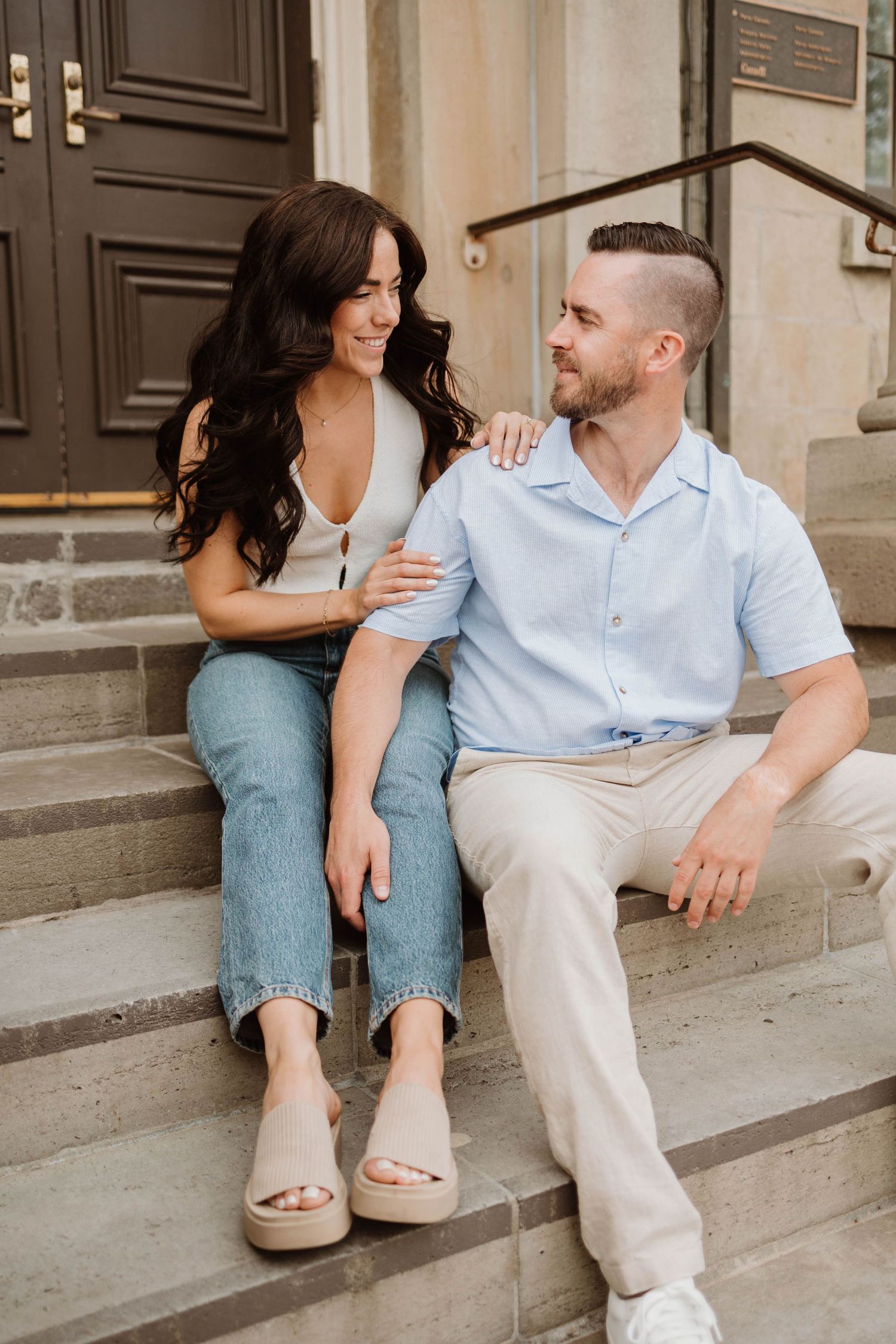 Couple sitting on stone steps smiling at each other, woman in jeans and white top, man in light blue shirt.