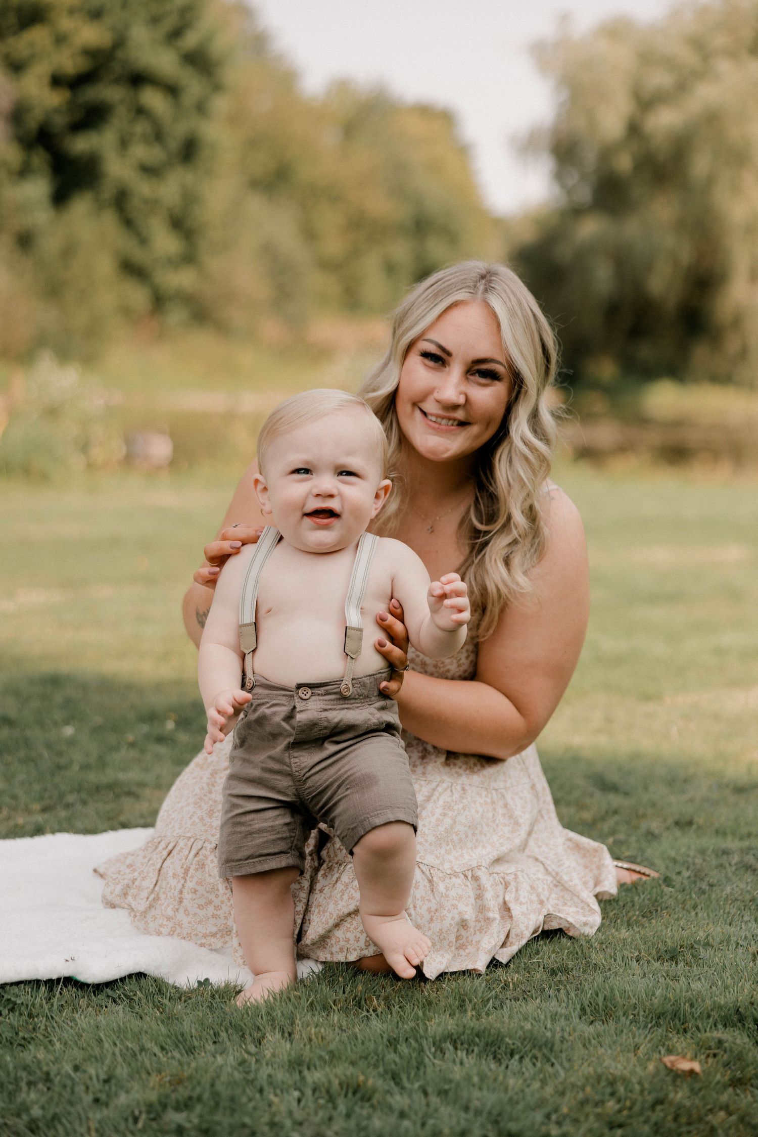 A smiling blonde mother sits on grass holding her laughing baby boy in suspenders during an outdoor photo session.