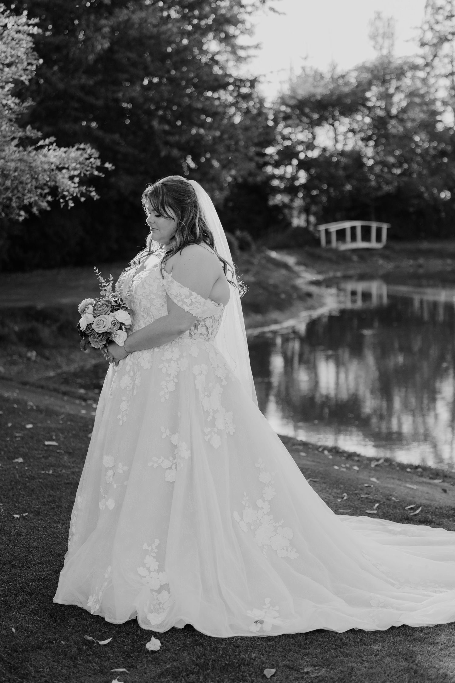 Black and white portrait of bride in floral lace gown with veil beside a serene pond.
