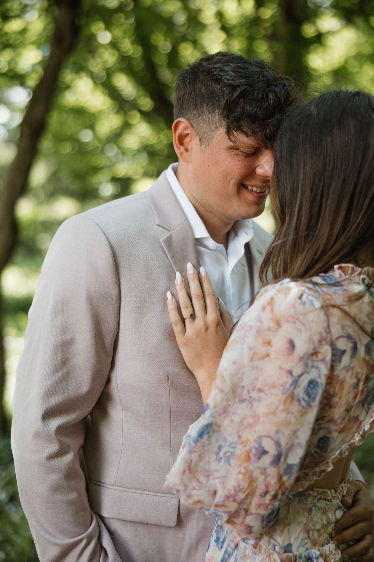 Man in beige suit smiling as his partner places her hand on his chest outdoors.