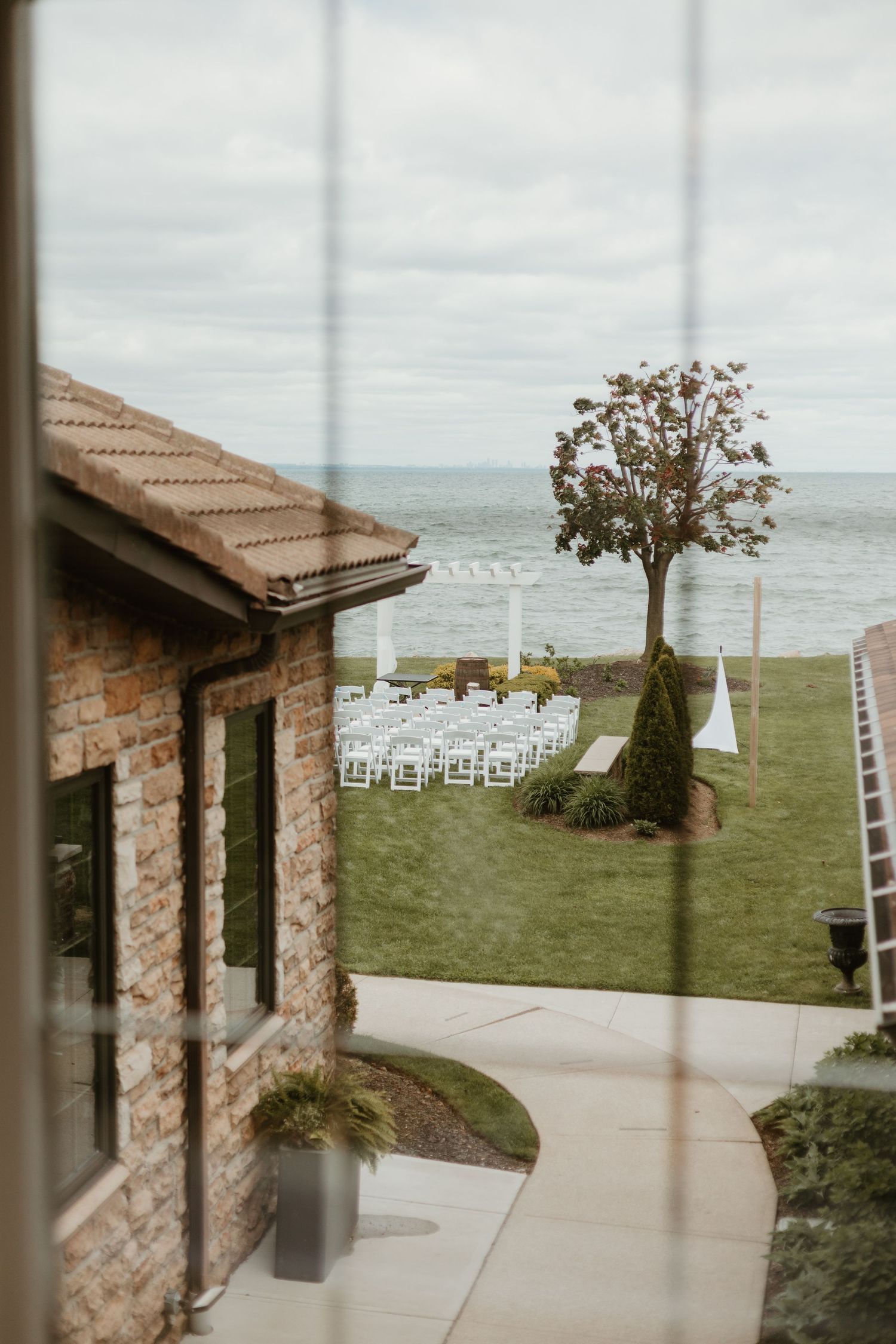 Elegant outdoor wedding ceremony setup with white chairs arranged by the ocean viewed through a window.