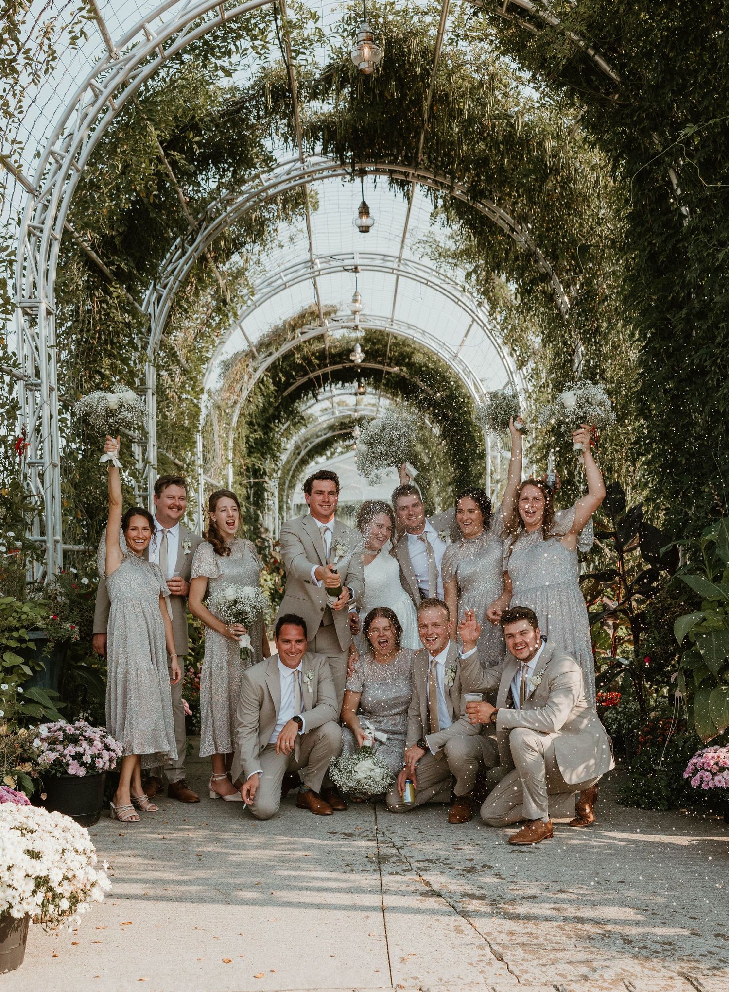 Wedding party celebrating under lush green arched greenhouse with bouquets raised.
