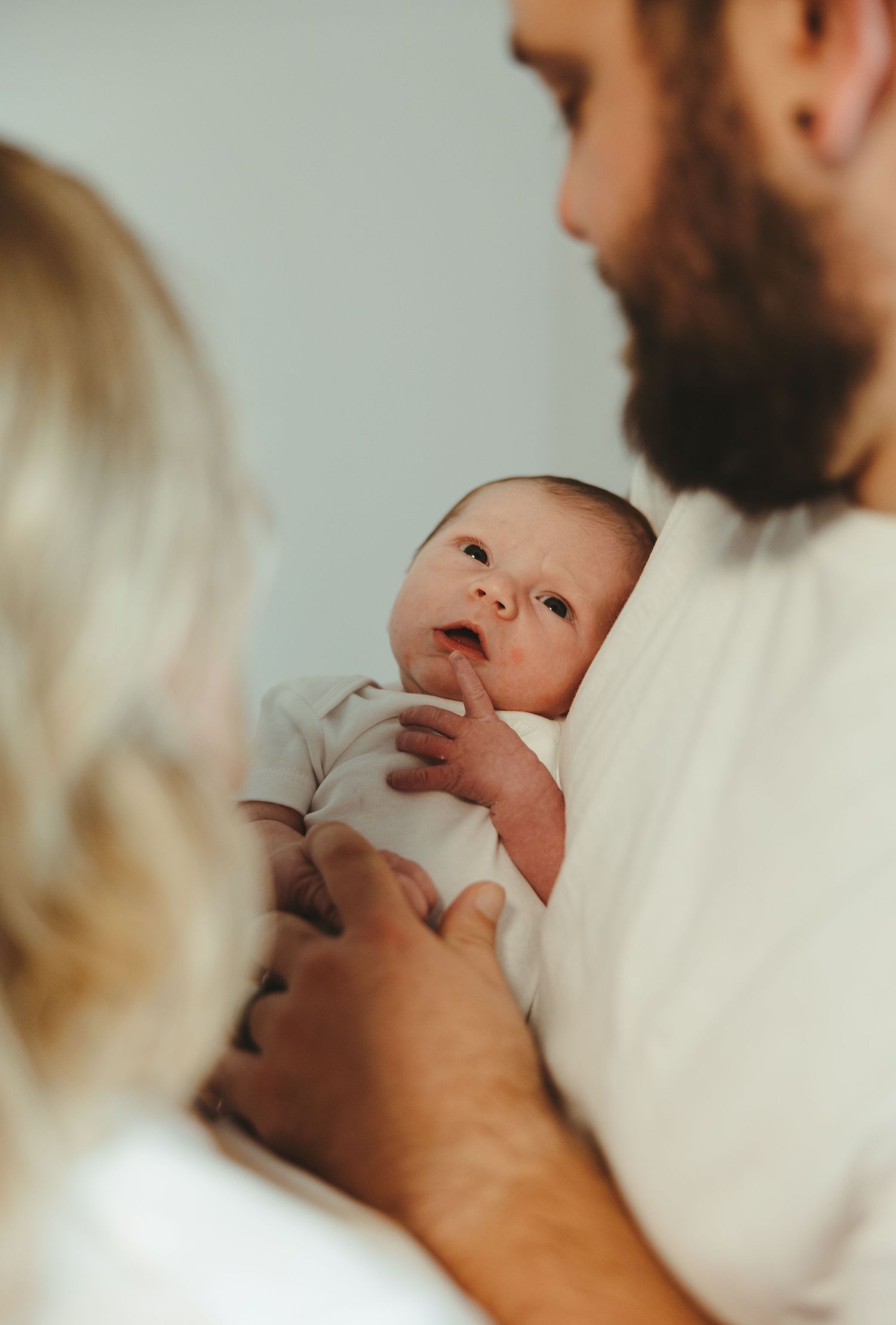Parents lovingly hold their alert newborn baby dressed in white onesie.