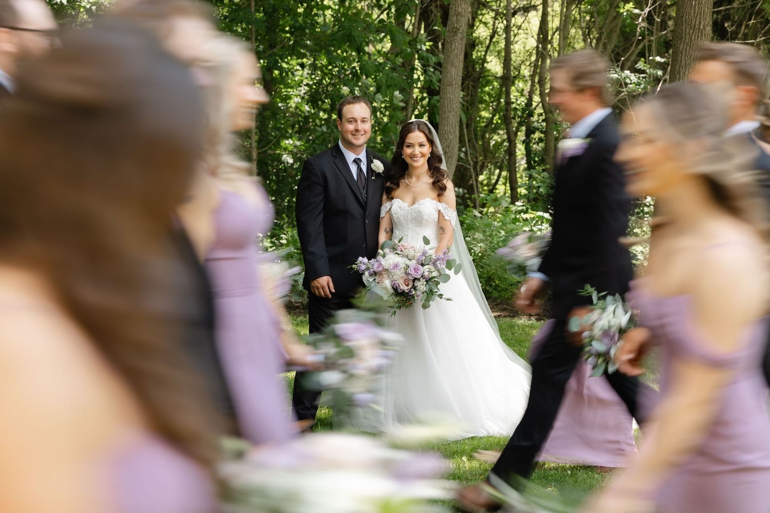 Smiling bride and groom stand together surrounded by bridesmaids in lavender dresses in wooded outdoor setting.