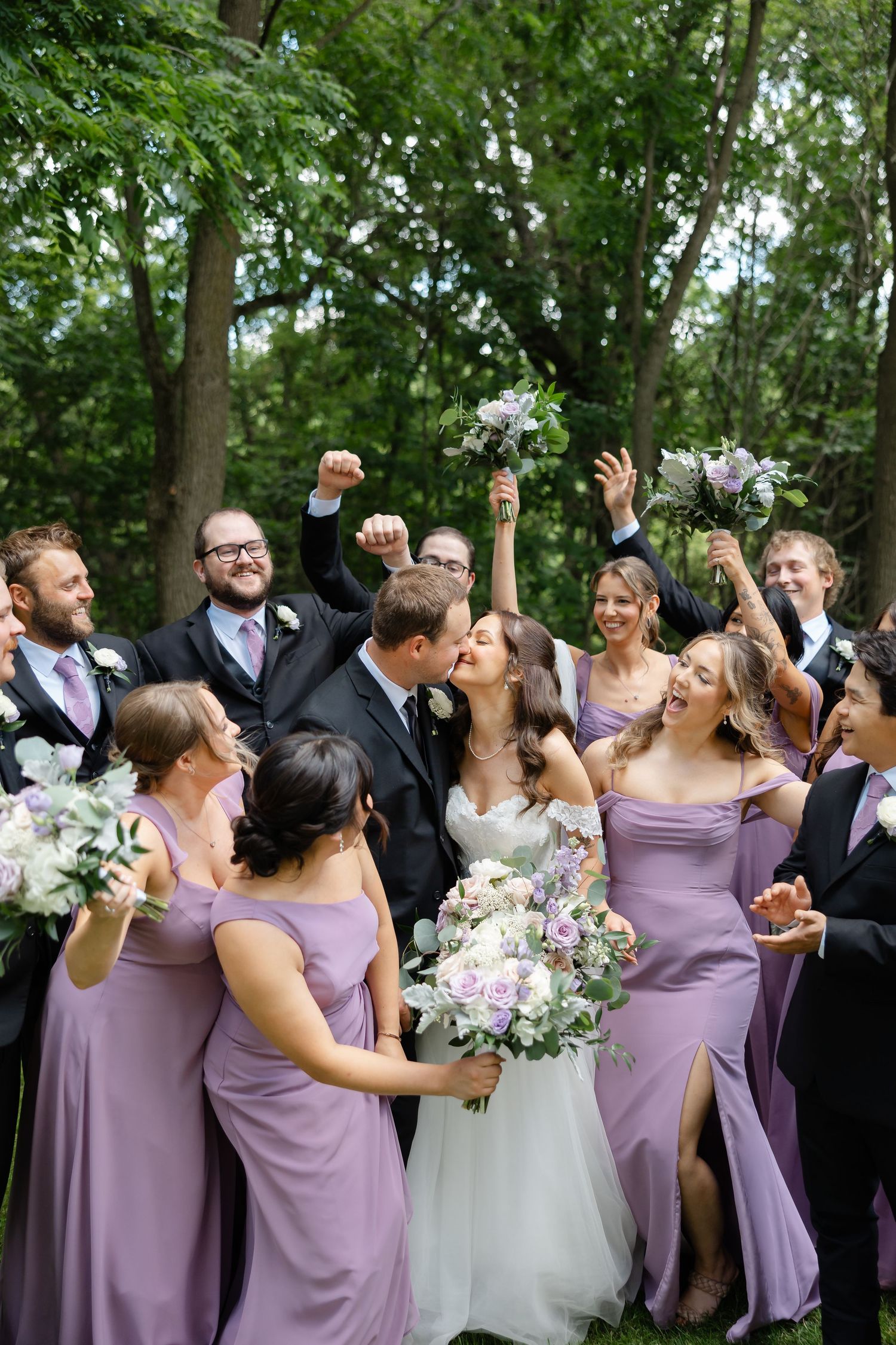 A joyful wedding party celebrates as the bride and groom kiss, surrounded by bridesmaids in lavender gowns outdoors.