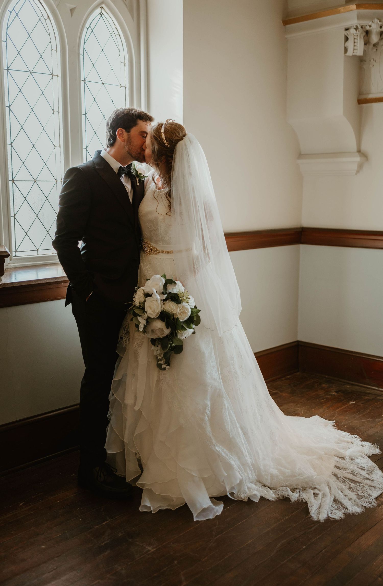 Bride and groom share a kiss by leaded glass window, bride in ruffled lace gown with long train.