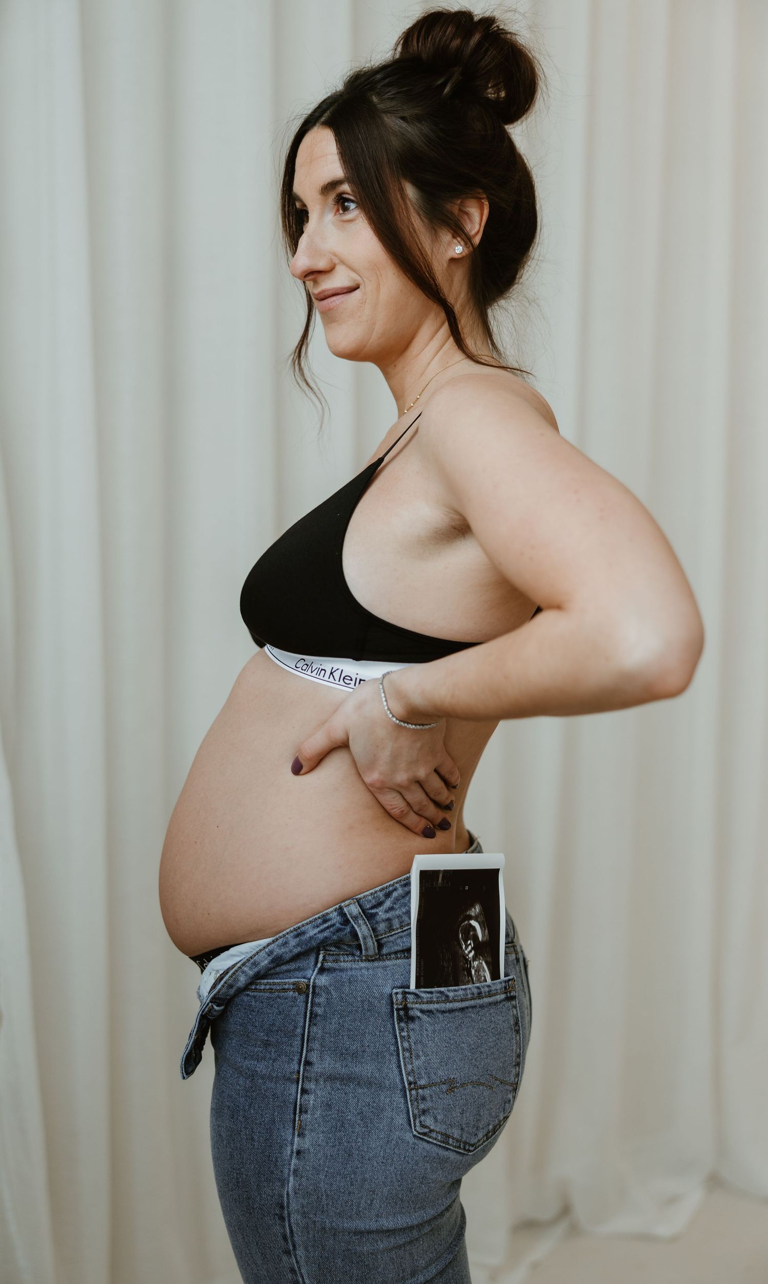 Pregnant woman in black bralette and jeans posing sideways with an ultrasound photo in her pocket.