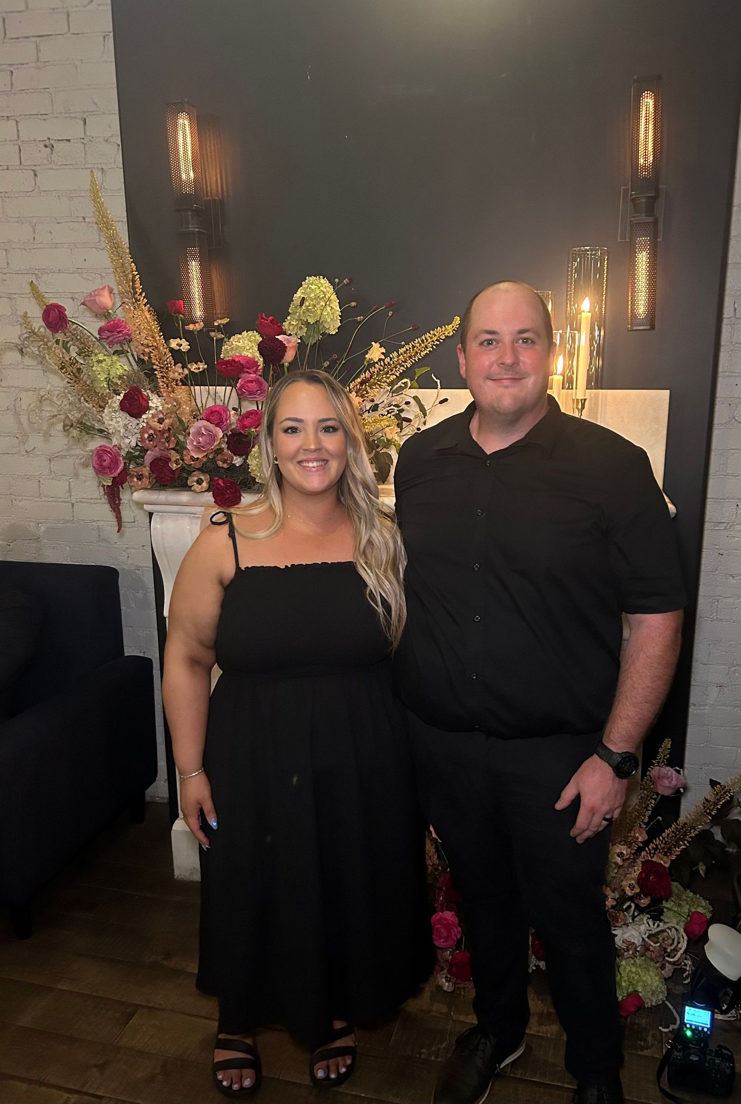 A couple dressed in black outfits smile together in front of a floral arrangement at an elegant venue.