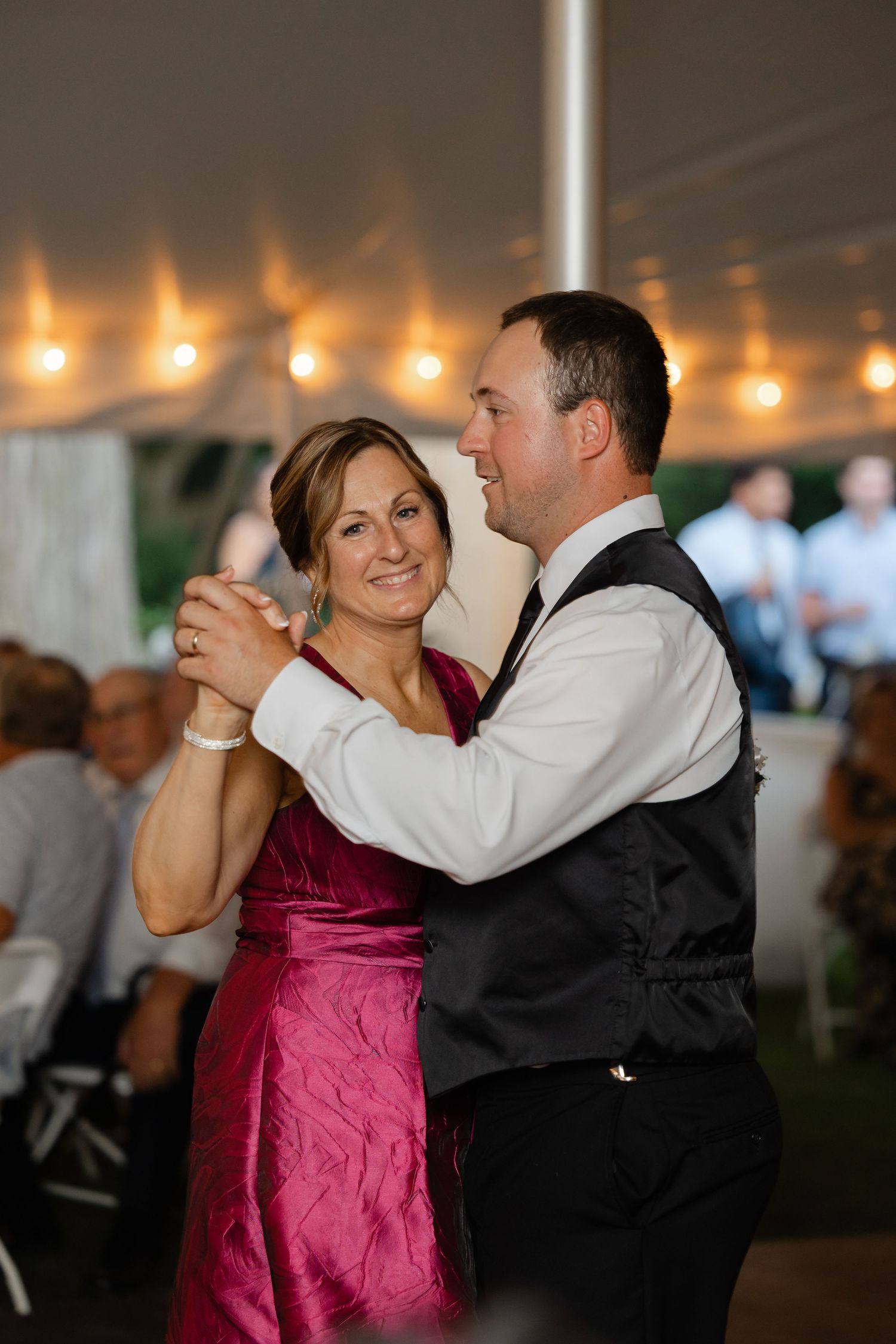 Mother and son share a tender dance under string lights at elegant tented wedding reception.