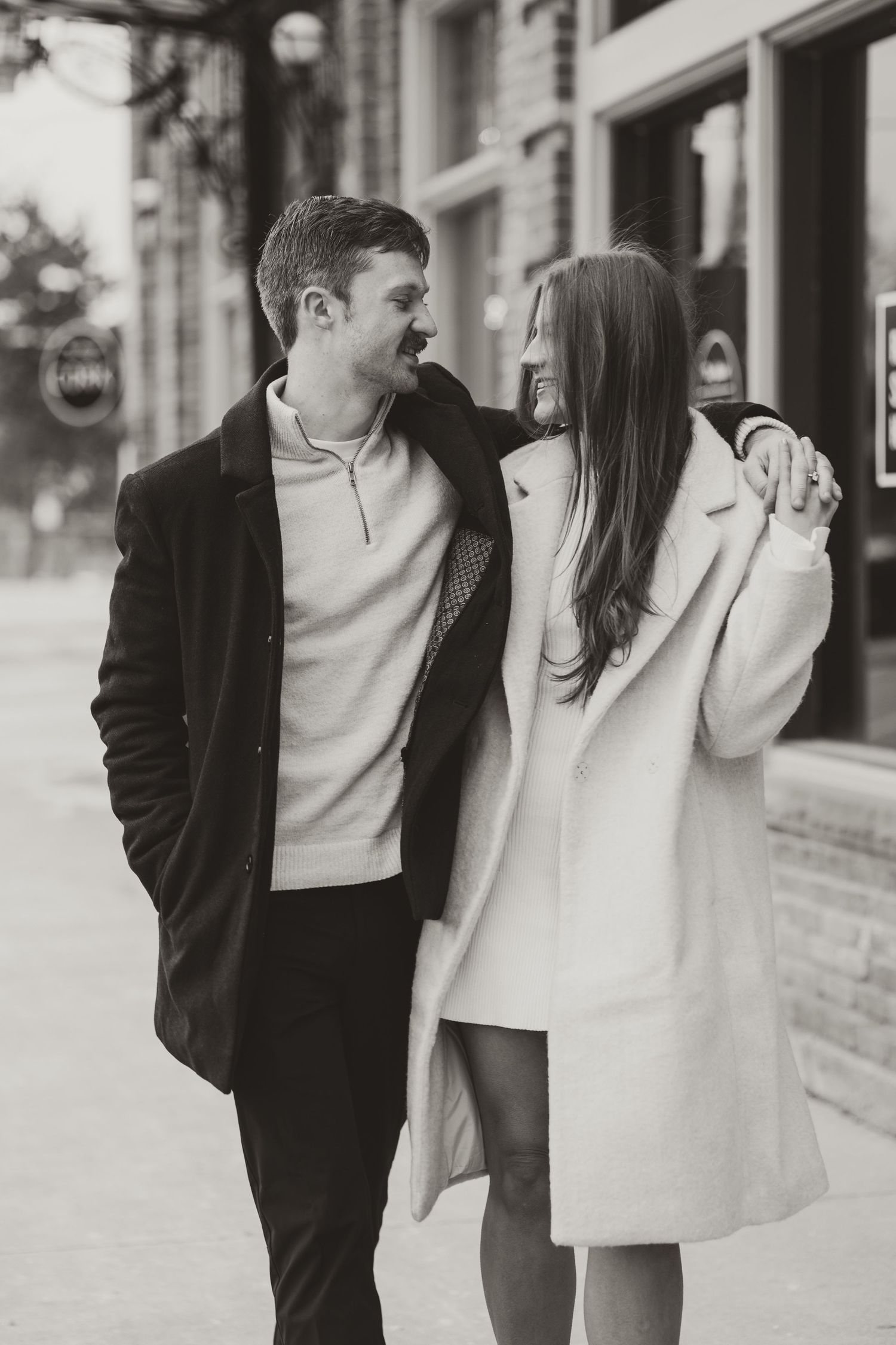 Black and white photo of a couple walking arm in arm on a city sidewalk, smiling at each other.