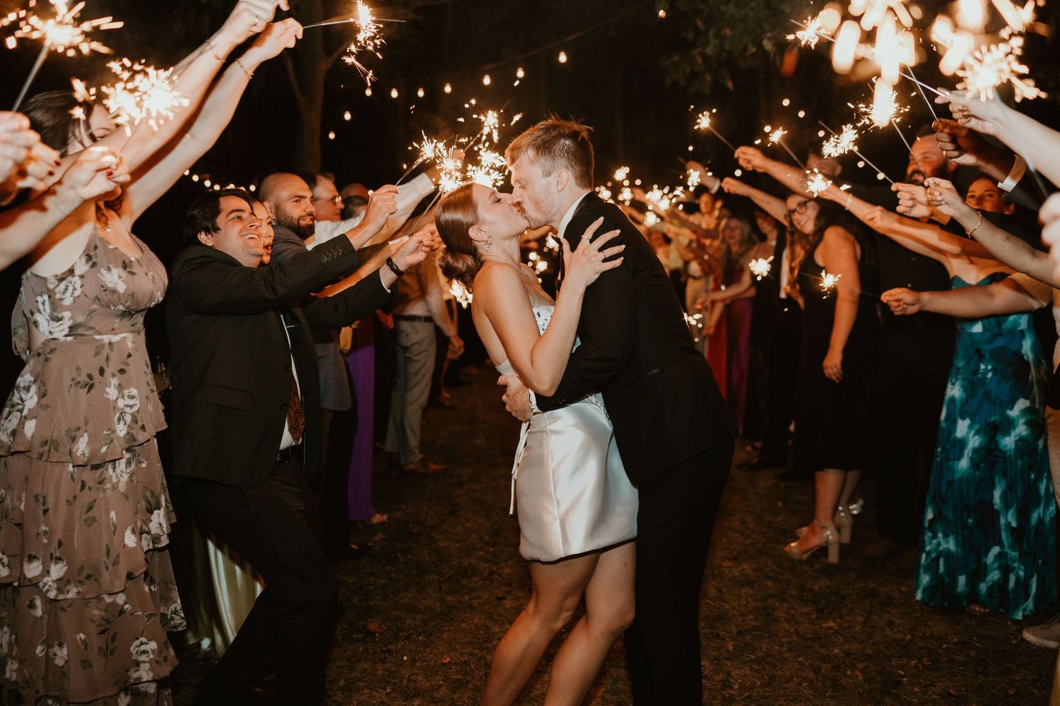 Bride and groom share a romantic kiss under a canopy of sparkling sparklers held by wedding guests at night.