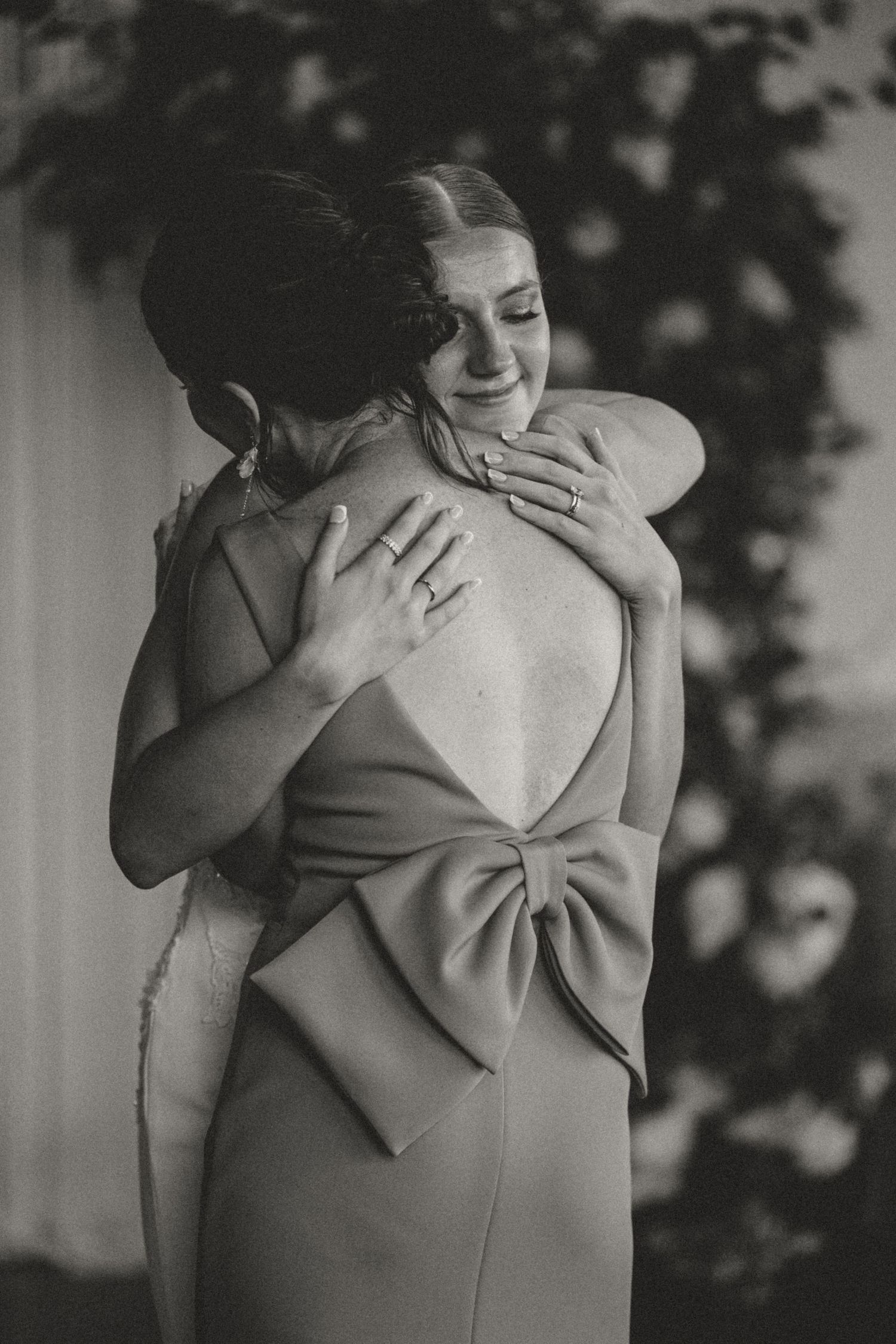 Black and white photo of bride and woman in bow-back dress sharing an emotional wedding hug.