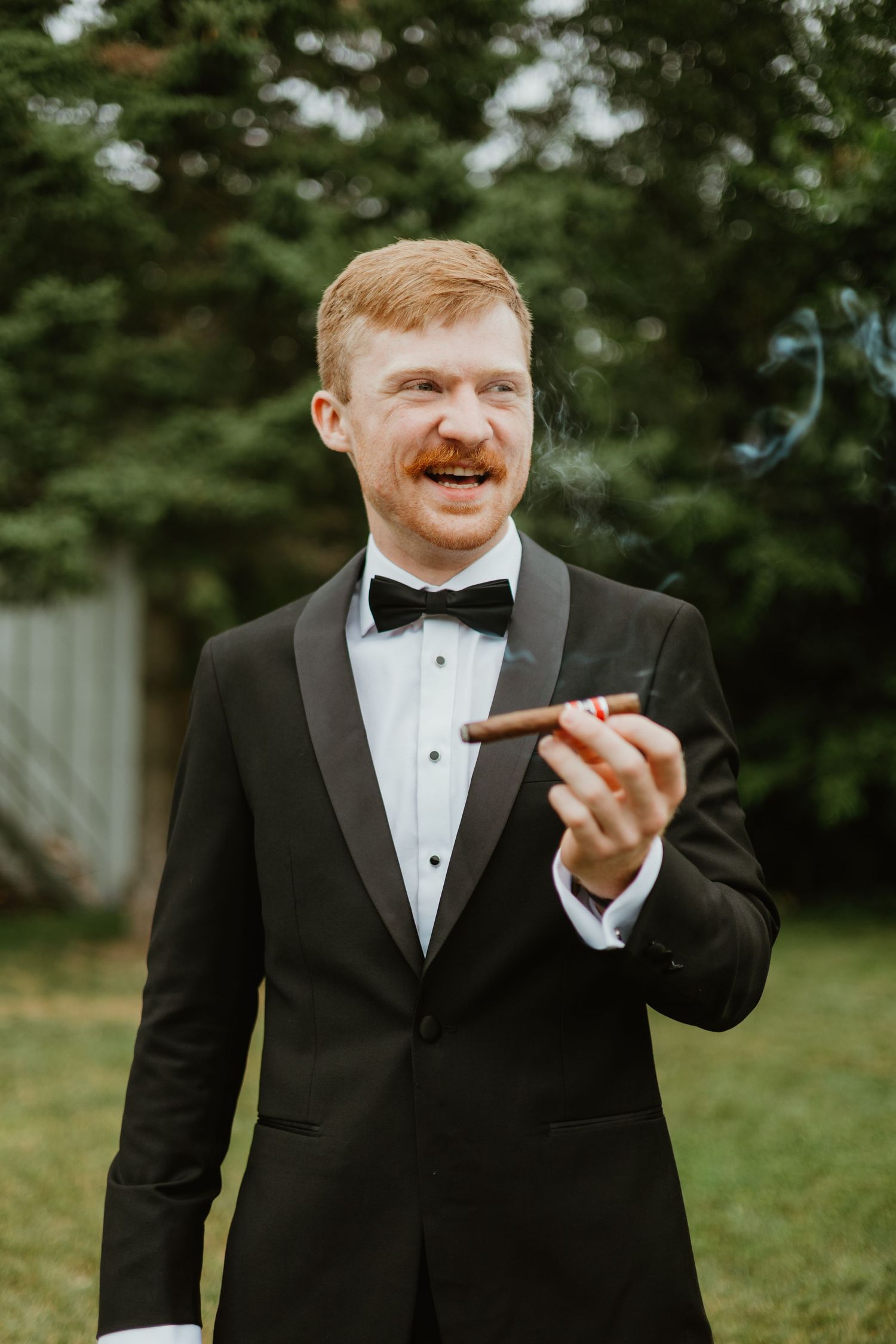 Dapper groom in black tuxedo with bow tie smiles while holding a cigar outdoors at wedding.