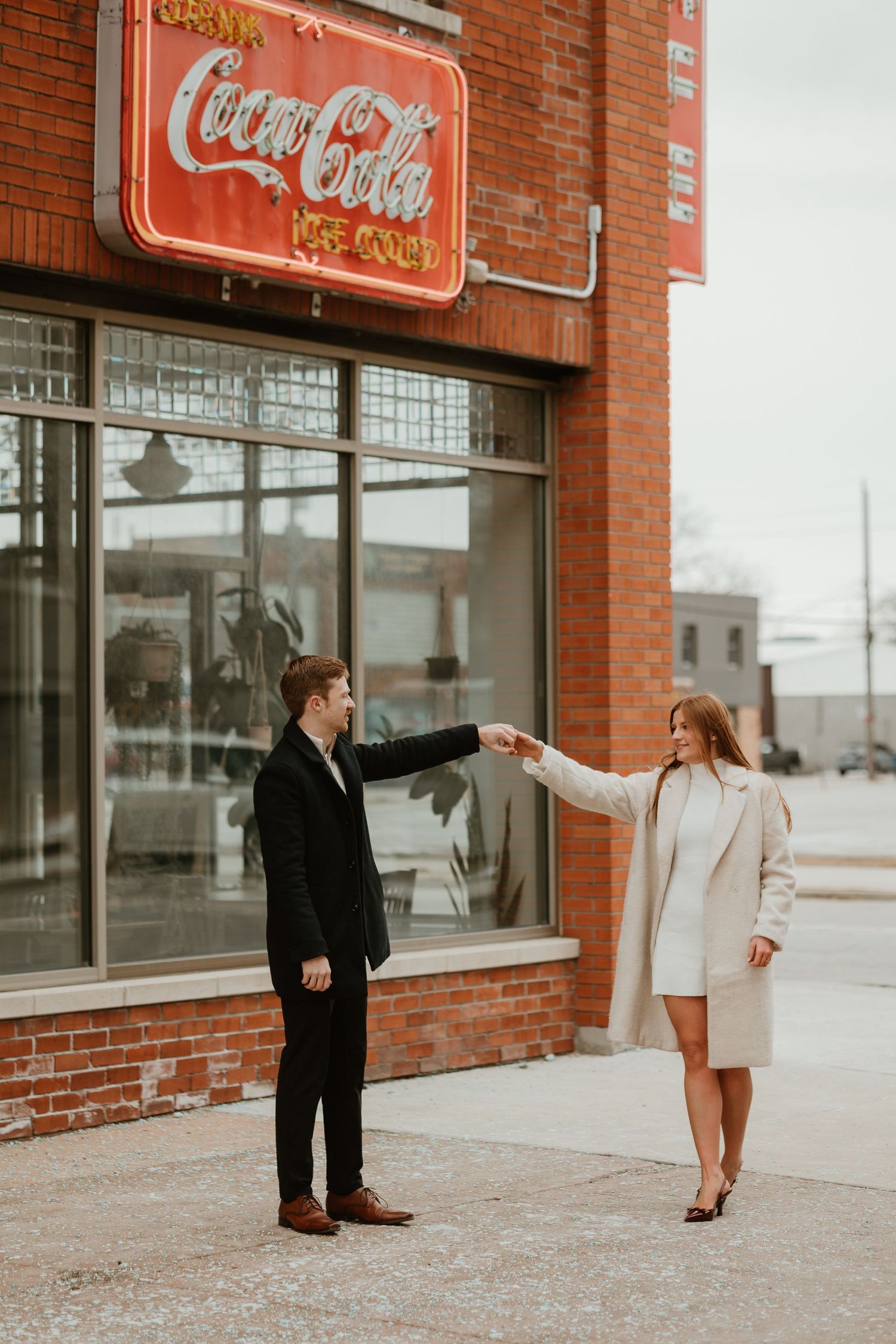 Couple holding hands in front of a vintage Coca-Cola neon sign on a brick building.