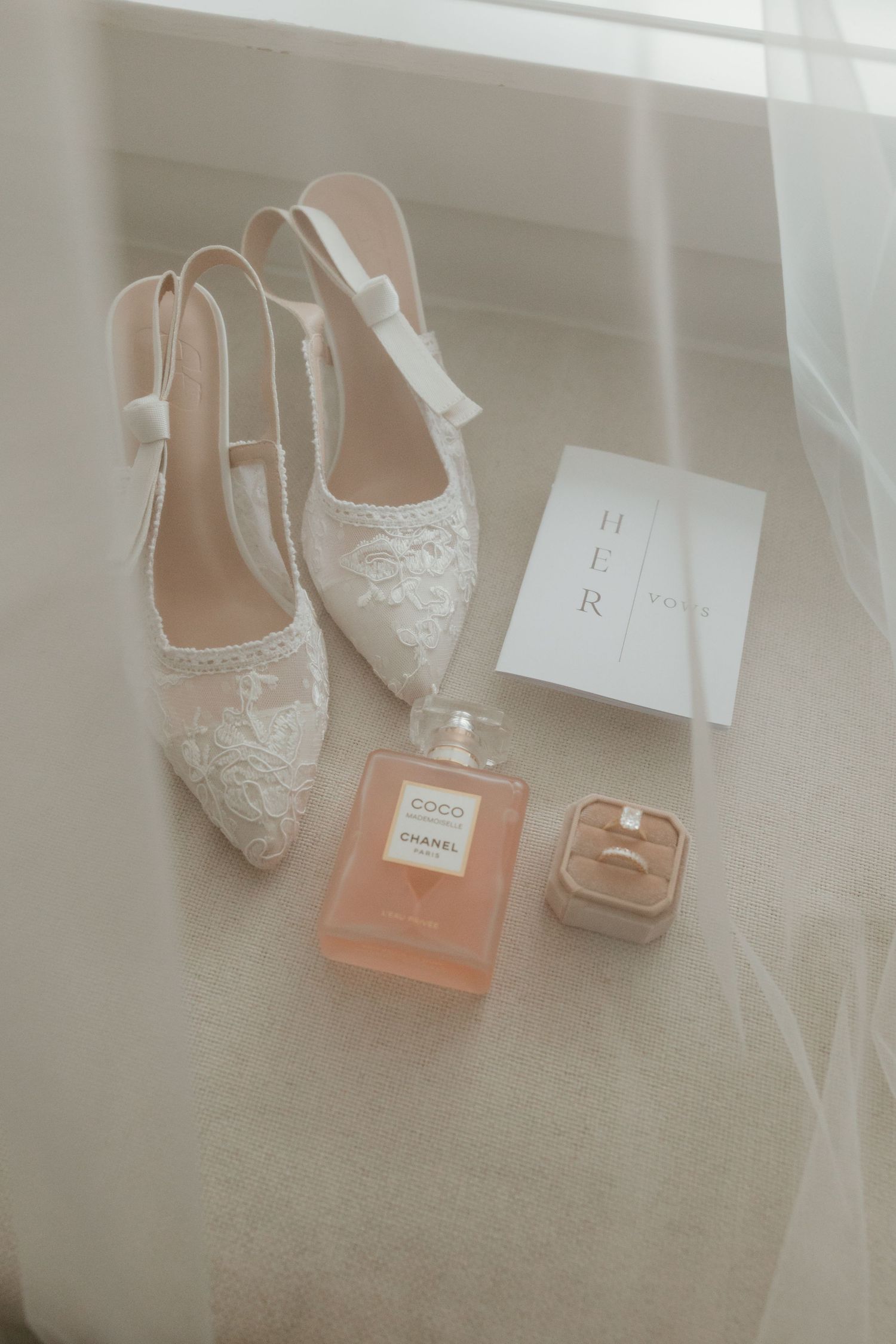 Bridal flat shoes with lace detail, Chanel Coco perfume, diamond ring, and wedding card styled on white surface.