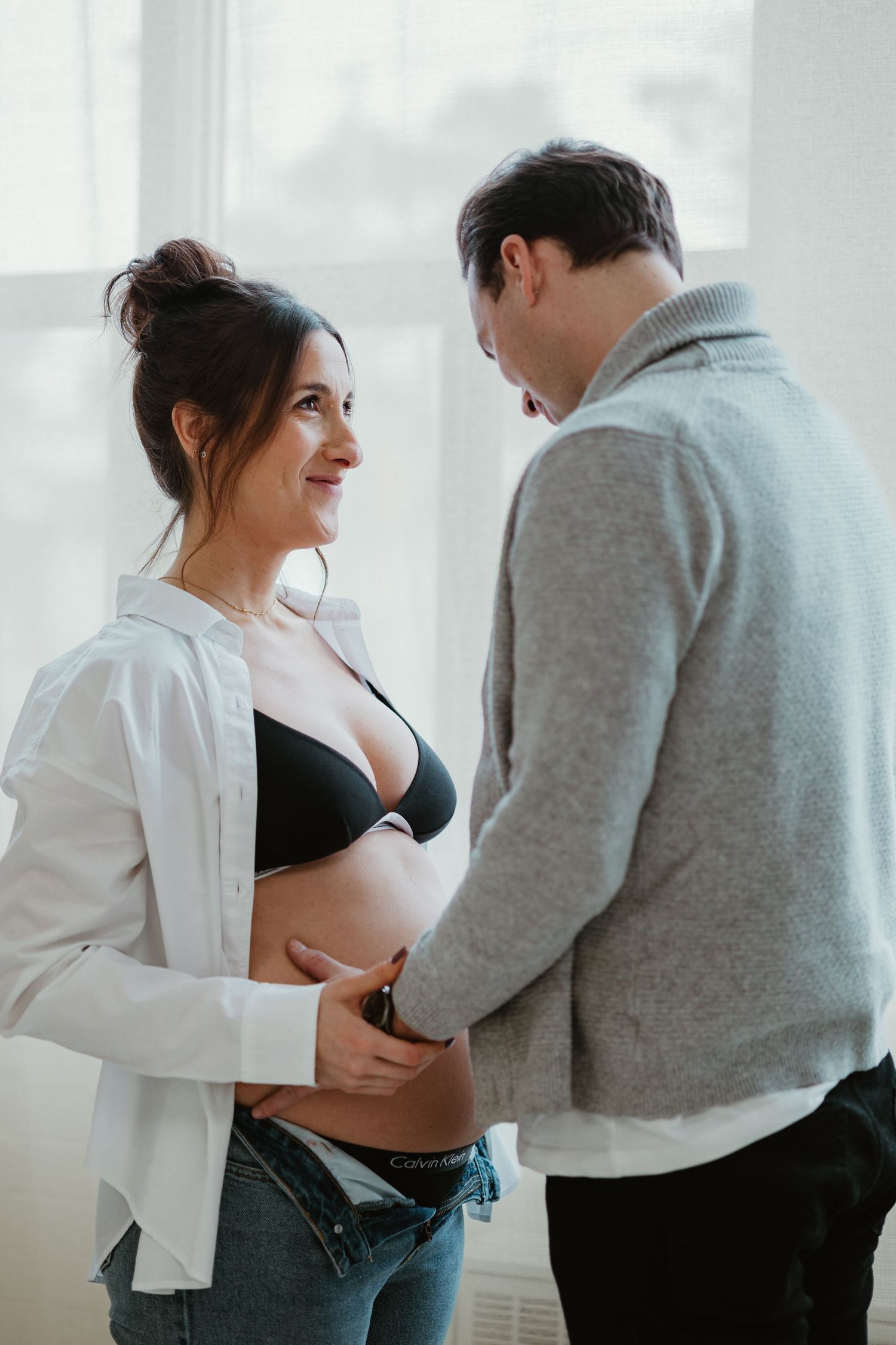 Pregnant woman in black bra smiling at her partner who gently touches her baby bump indoors.