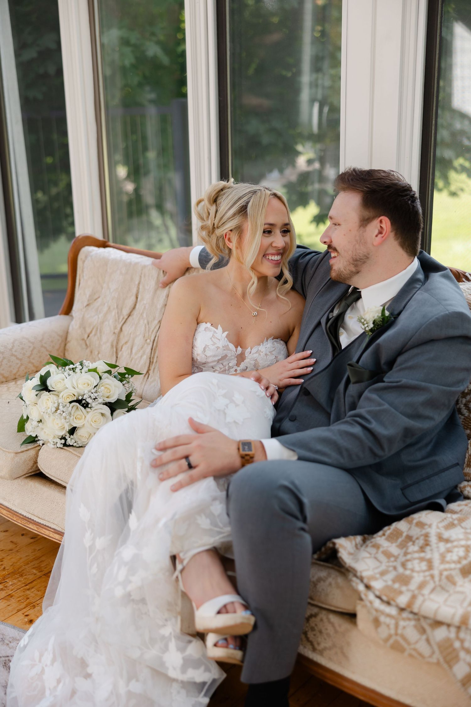 Smiling bride in a lace gown and groom in a gray suit embrace on a cream sofa beside a white floral wedding bouquet.