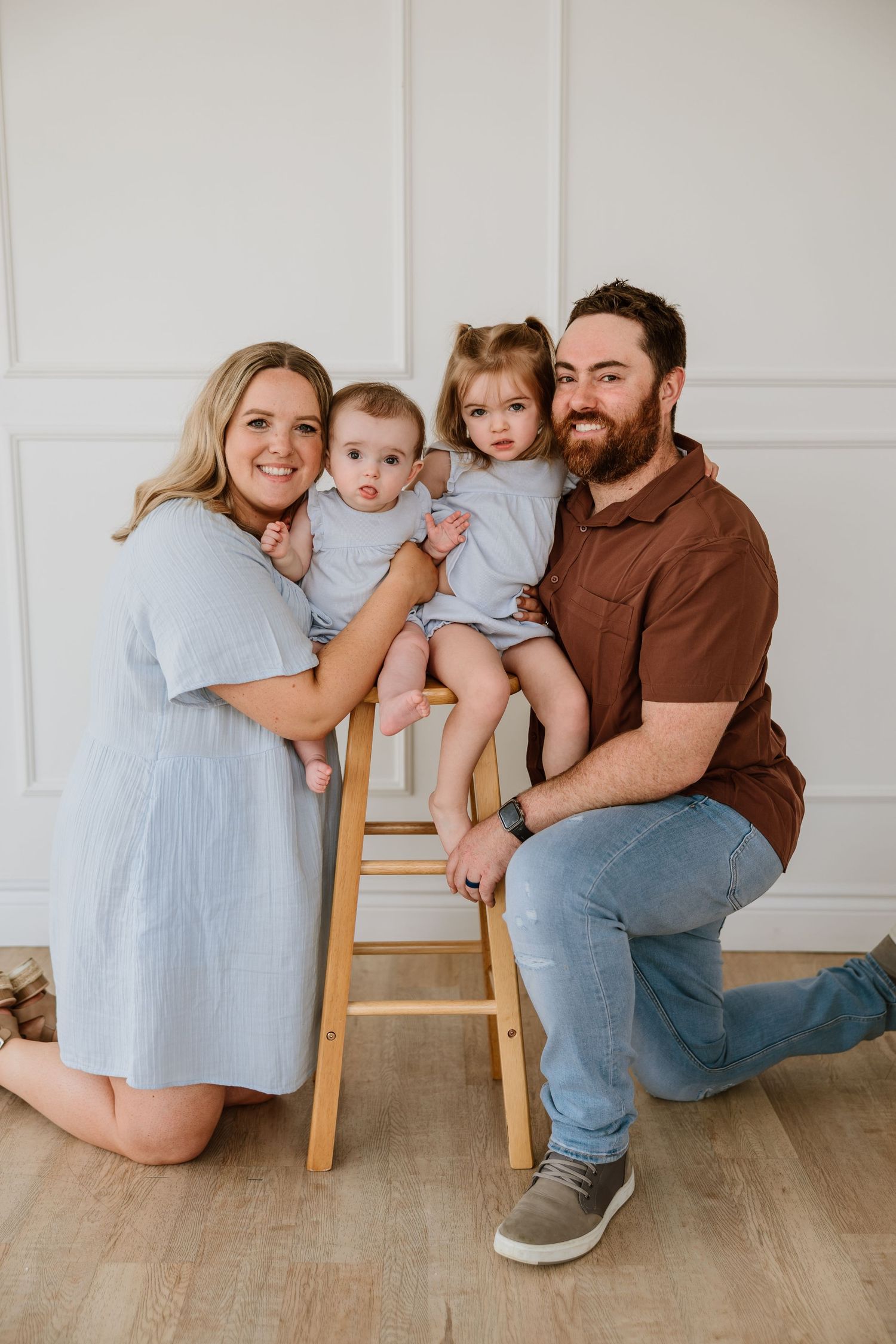 Happy family of four poses together in a bright studio with two young children on a wooden stool.