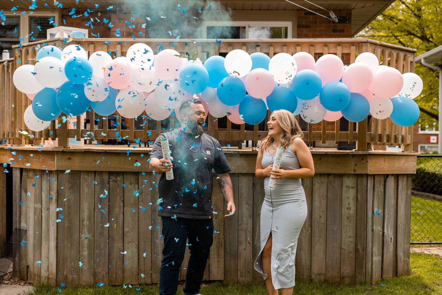 Couple laughing at outdoor gender reveal with blue confetti and pink and blue balloon arch.